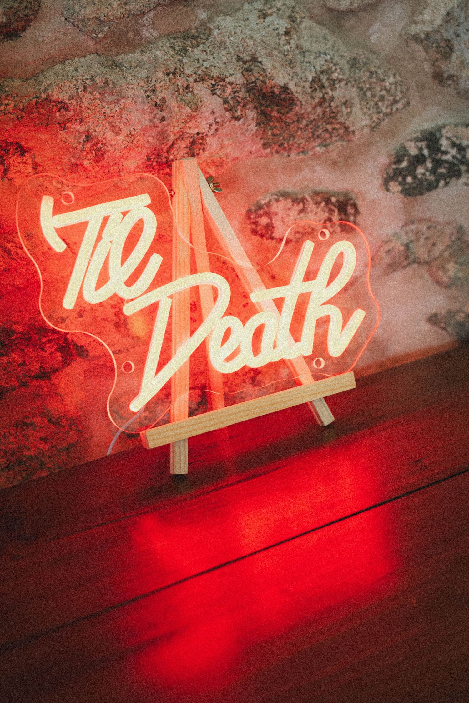 Glowing red neon sign reading Til Death displayed on wooden easel against rustic stone wall at wedding venue.