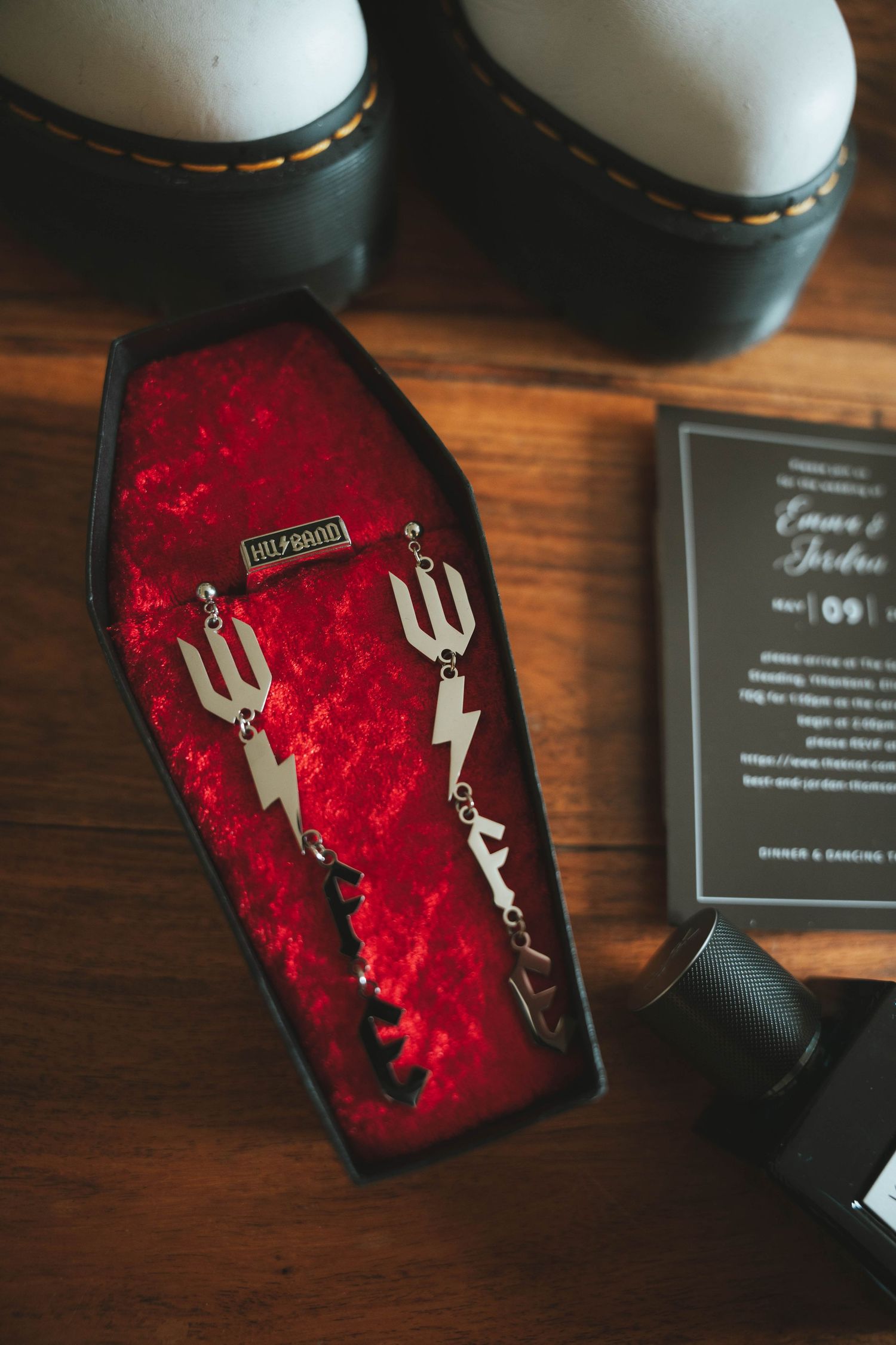 Gothic coffin jewelry box with red velvet interior displaying silver trident earrings, beside Dr. Martens boots and wedding invitation.