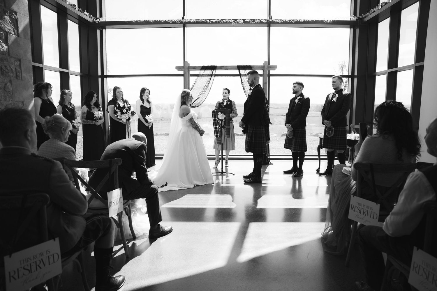 Black and white wedding ceremony photo with bride, groom, and officiant before large windows.