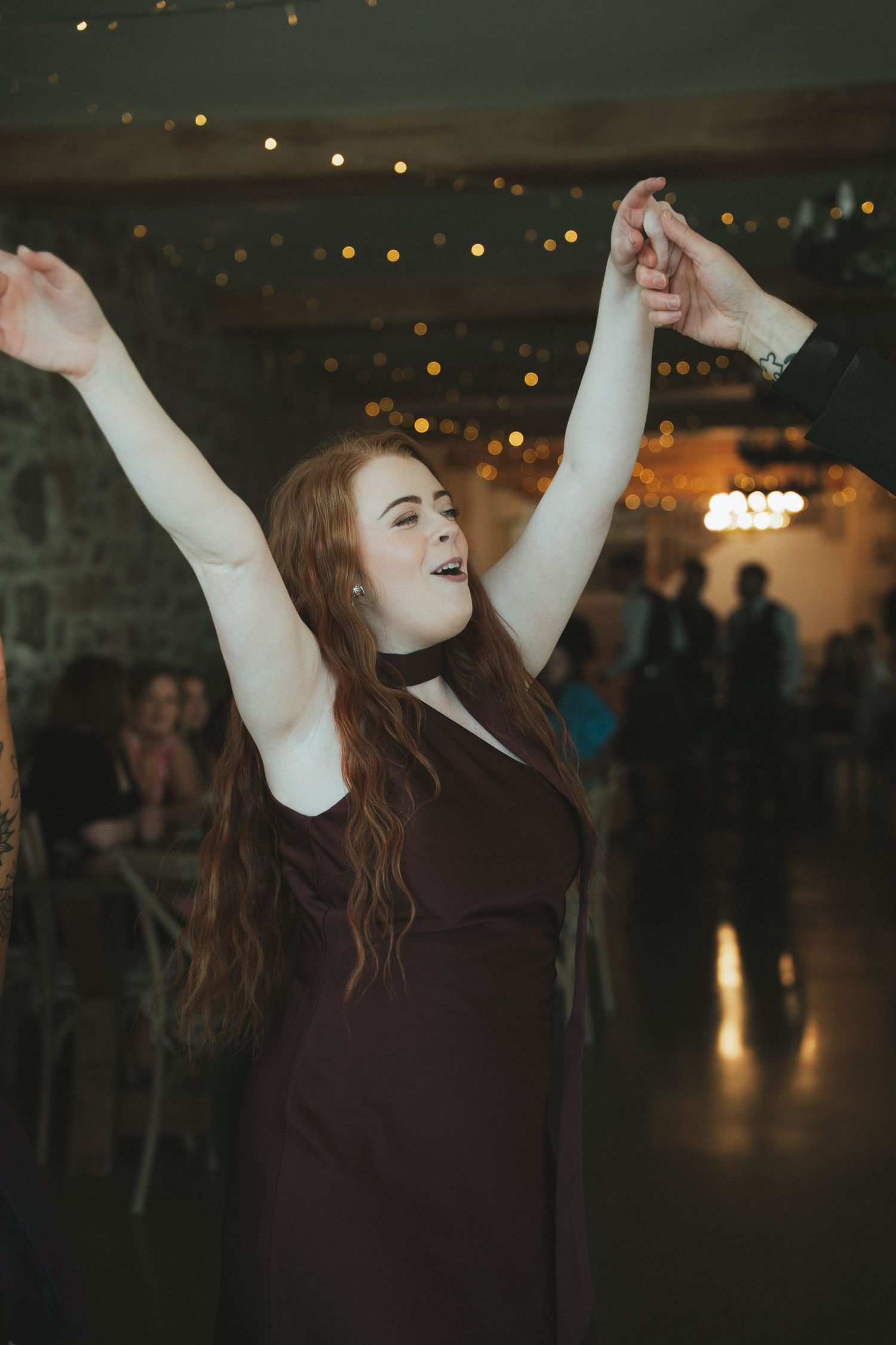 Red-haired woman in dark dress dances joyfully with arms raised at wedding reception party.