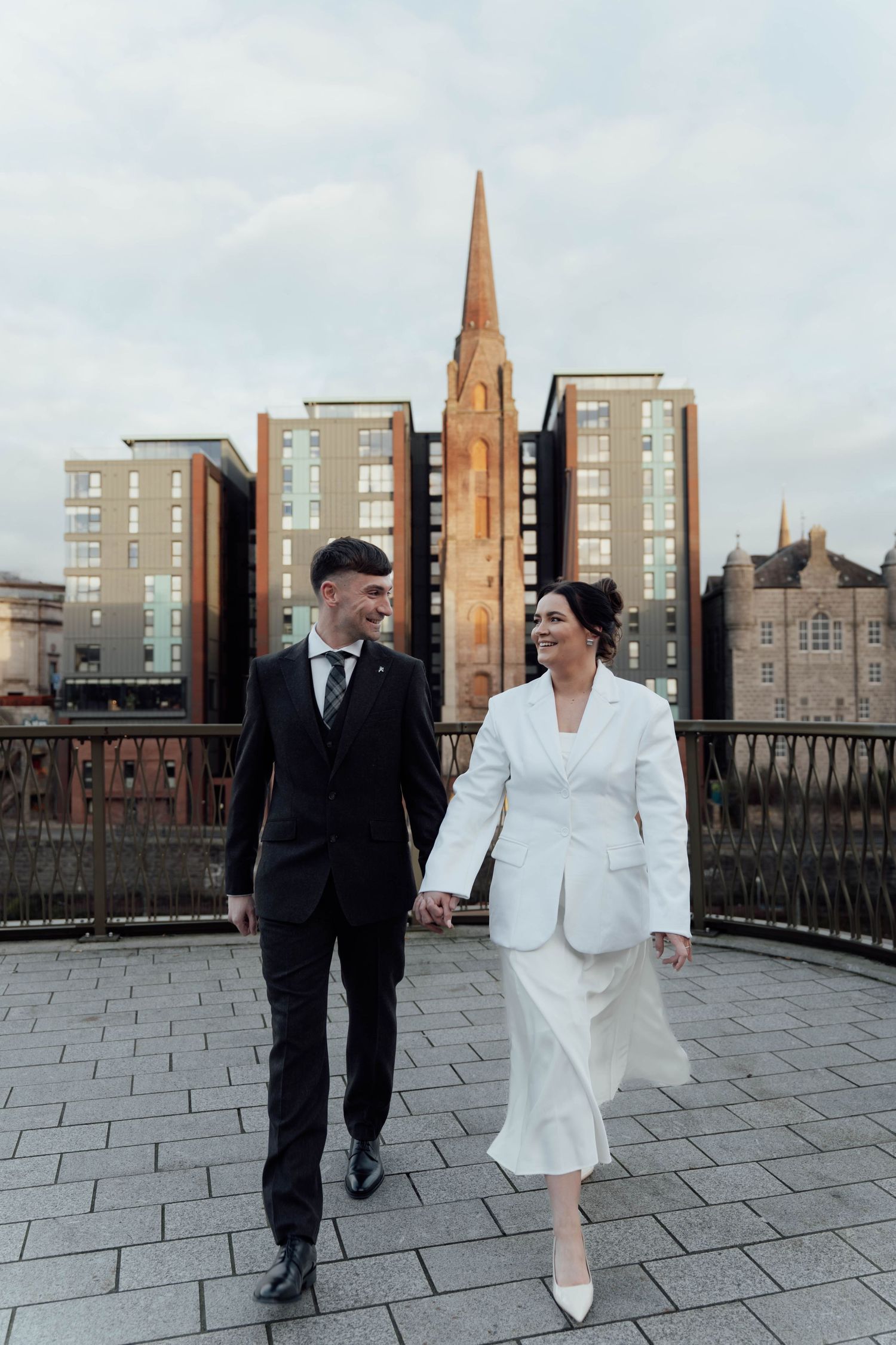 A couple walks hand in hand on a bridge with a church spire and modern buildings in the background.