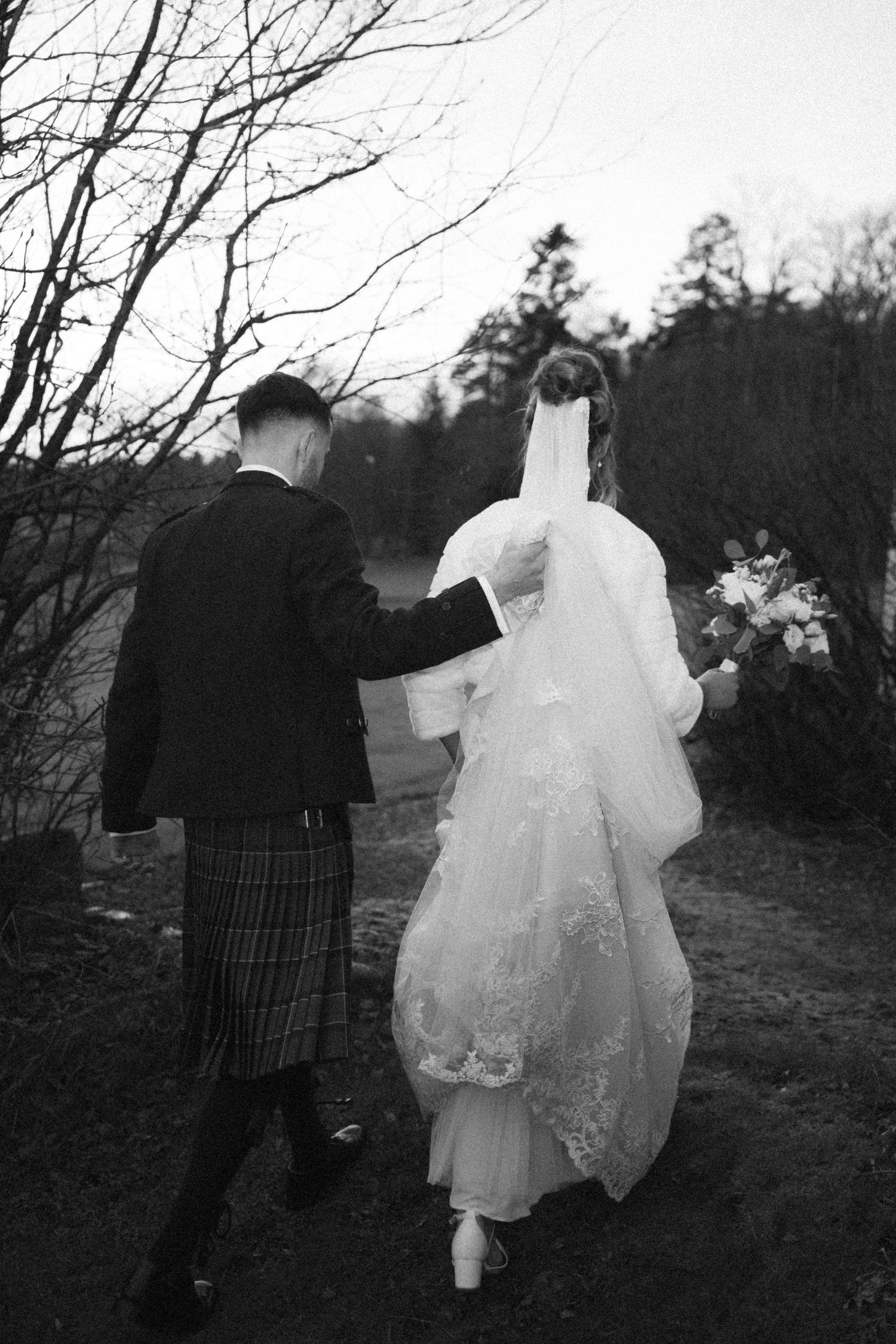 A couple walks together on a winter path, with the groom in a traditional kilt and bride in a flowing white dress.