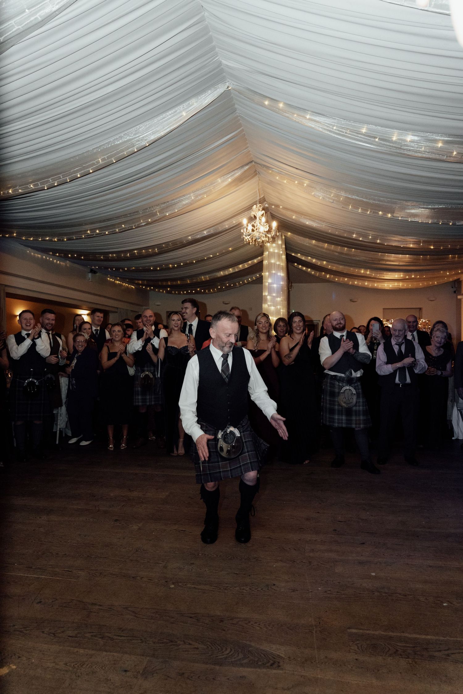 Wedding guests watch as dancers perform in a tent-draped reception venue with string lights.