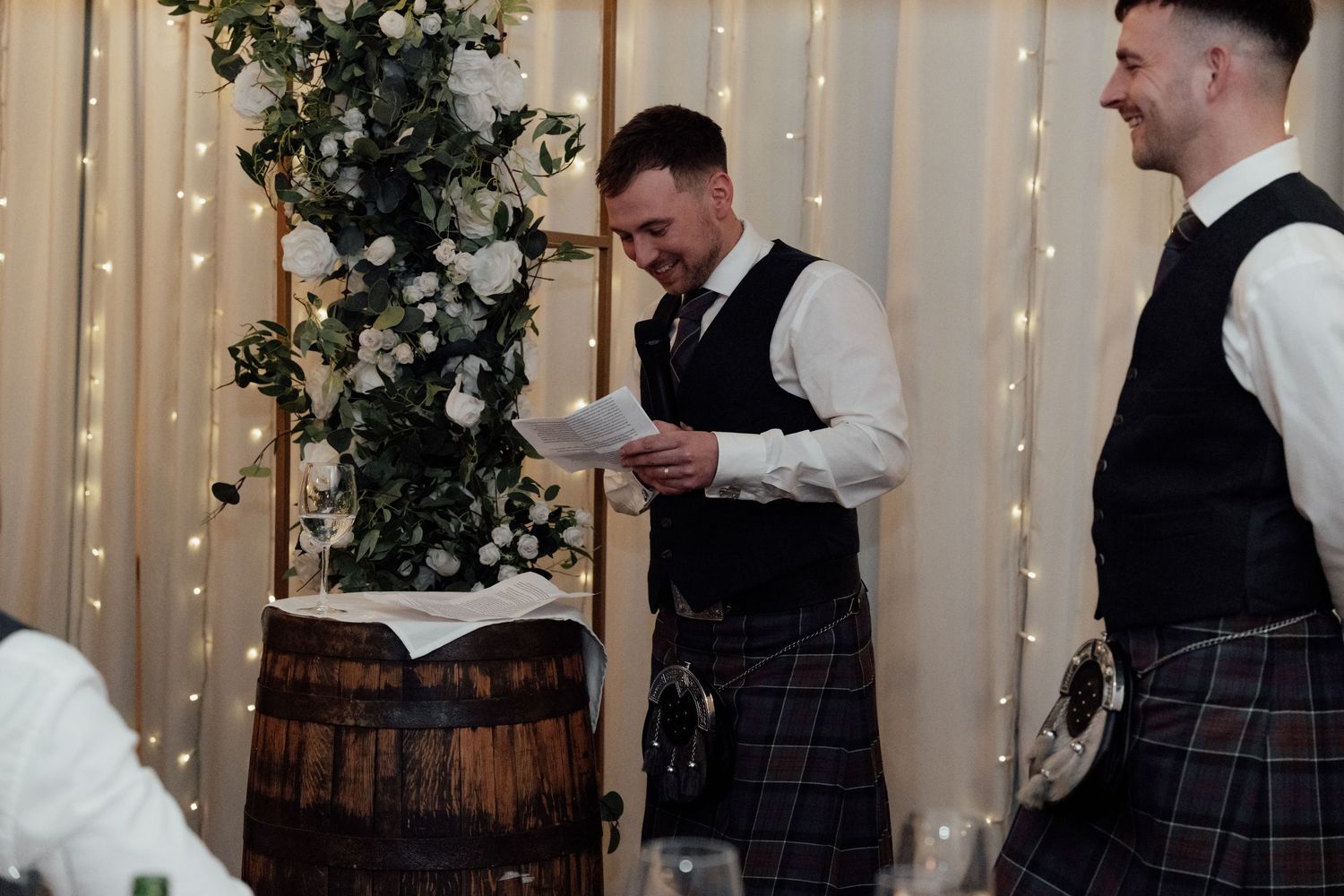 Groomsmen in traditional kilts standing near a rustic wine barrel and floral decorations.