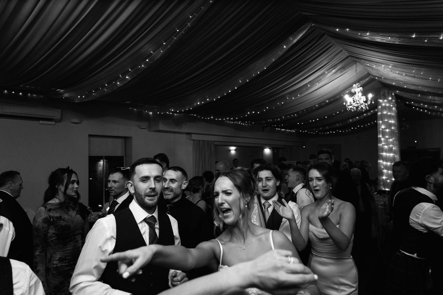 Wedding guests dancing and celebrating under elegant draped ceiling decorations.
