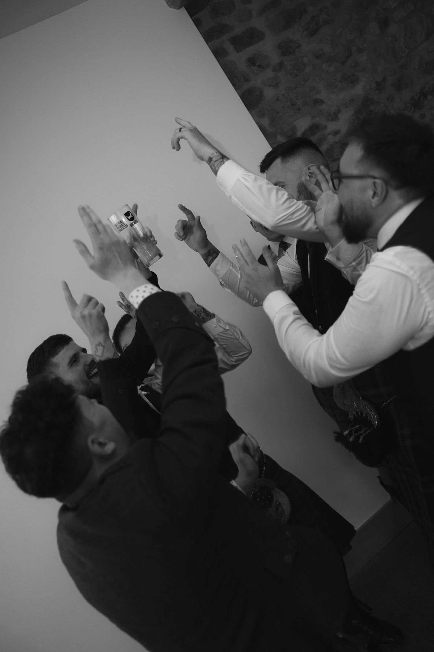 Black and white photo of groomsmen in kilts celebrating with hands raised at reception.