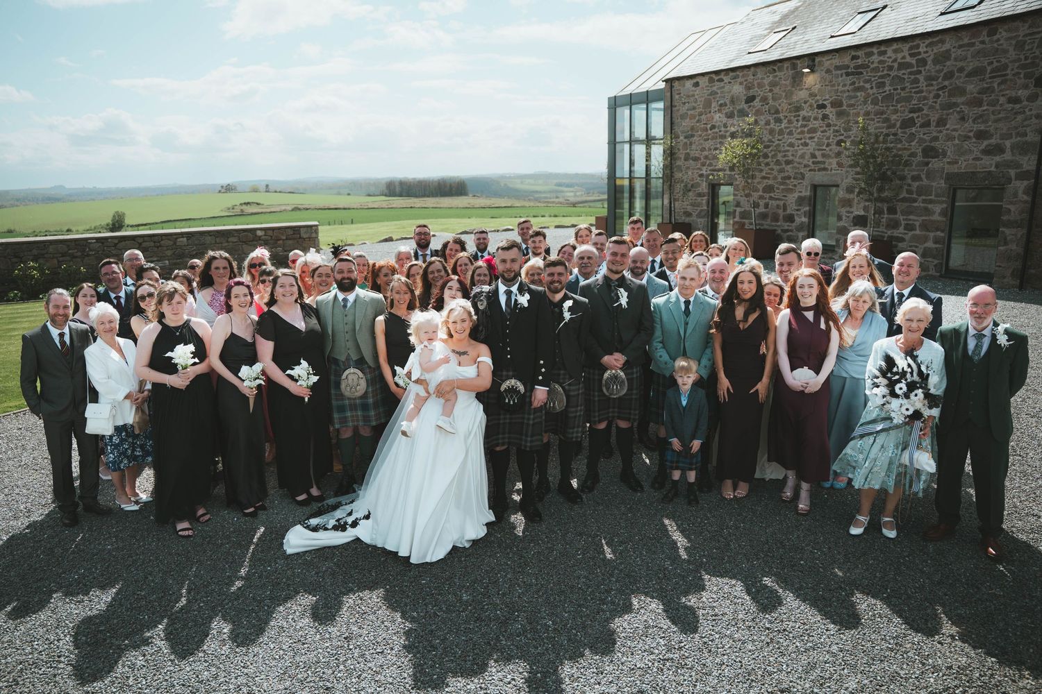 Large wedding party group photo outside rustic stone venue under blue sky in Scottish countryside.