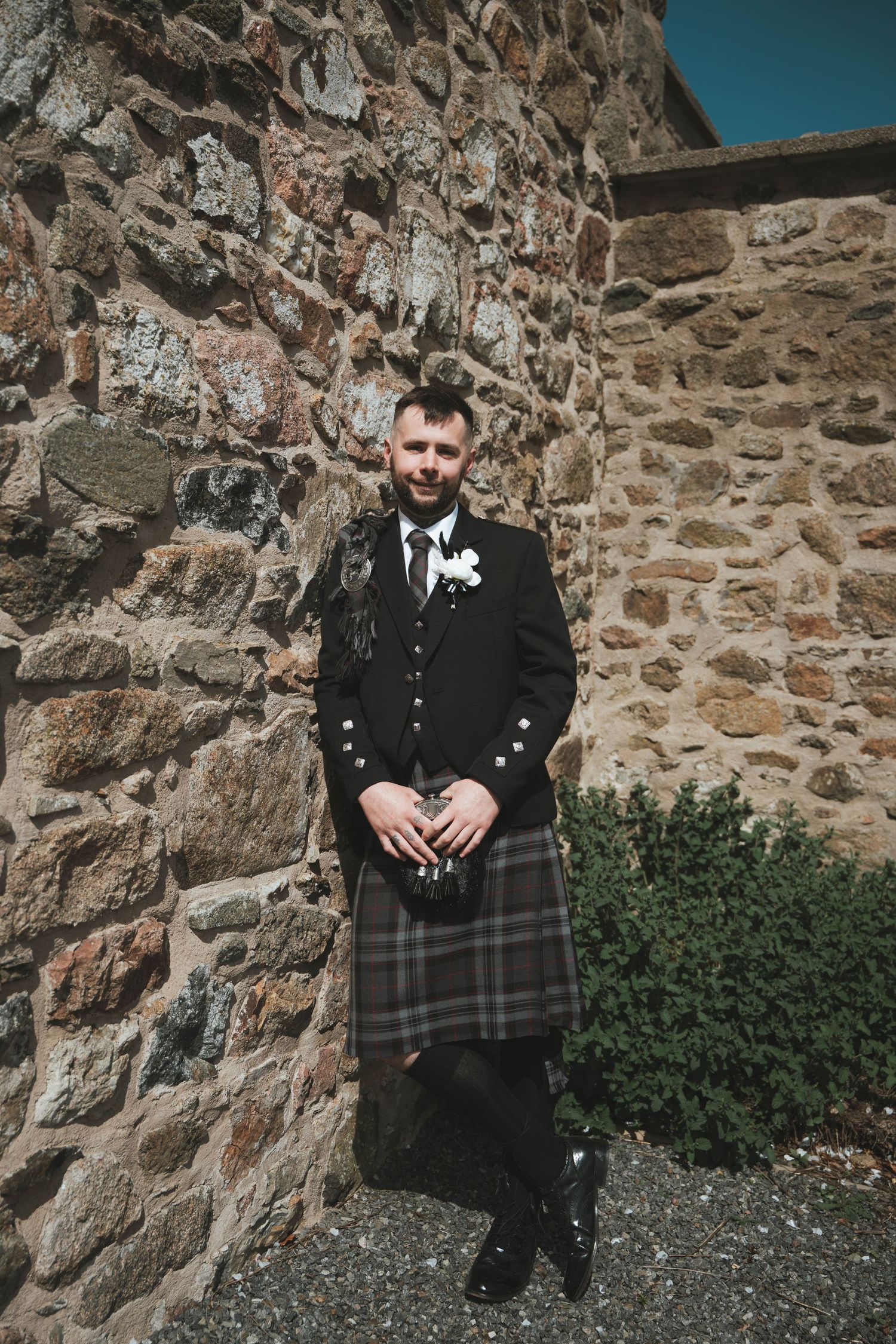 Groom in traditional Scottish kilt and black jacket with boutonniere stands against rustic stone wall.
