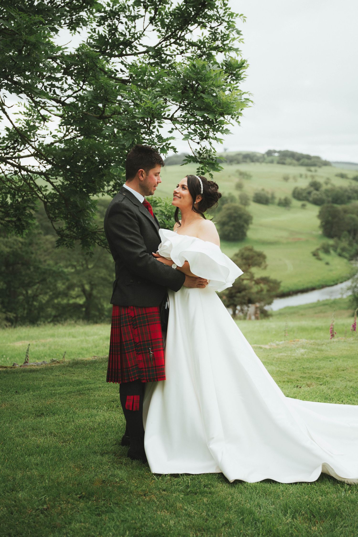 Bride in white gown and groom in red tartan kilt embrace outdoors with lush green Scottish countryside backdrop.