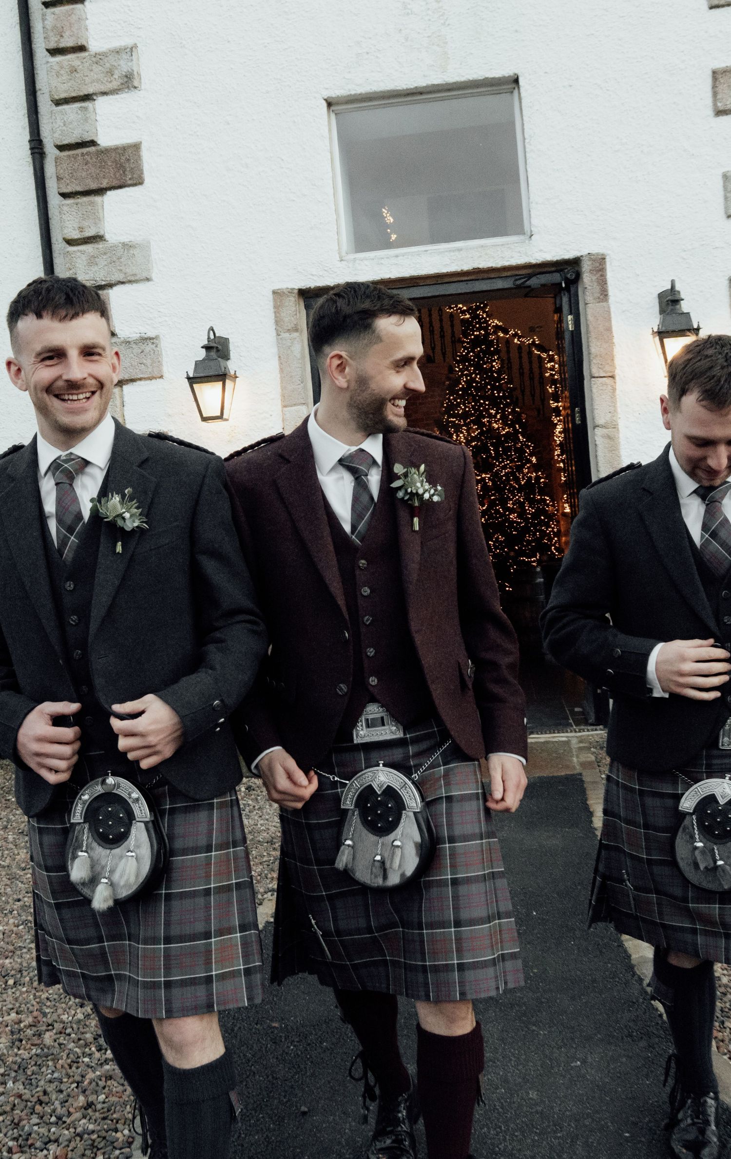 Groomsmen wearing traditional Scottish kilts and formal jackets pose together outside a white building.