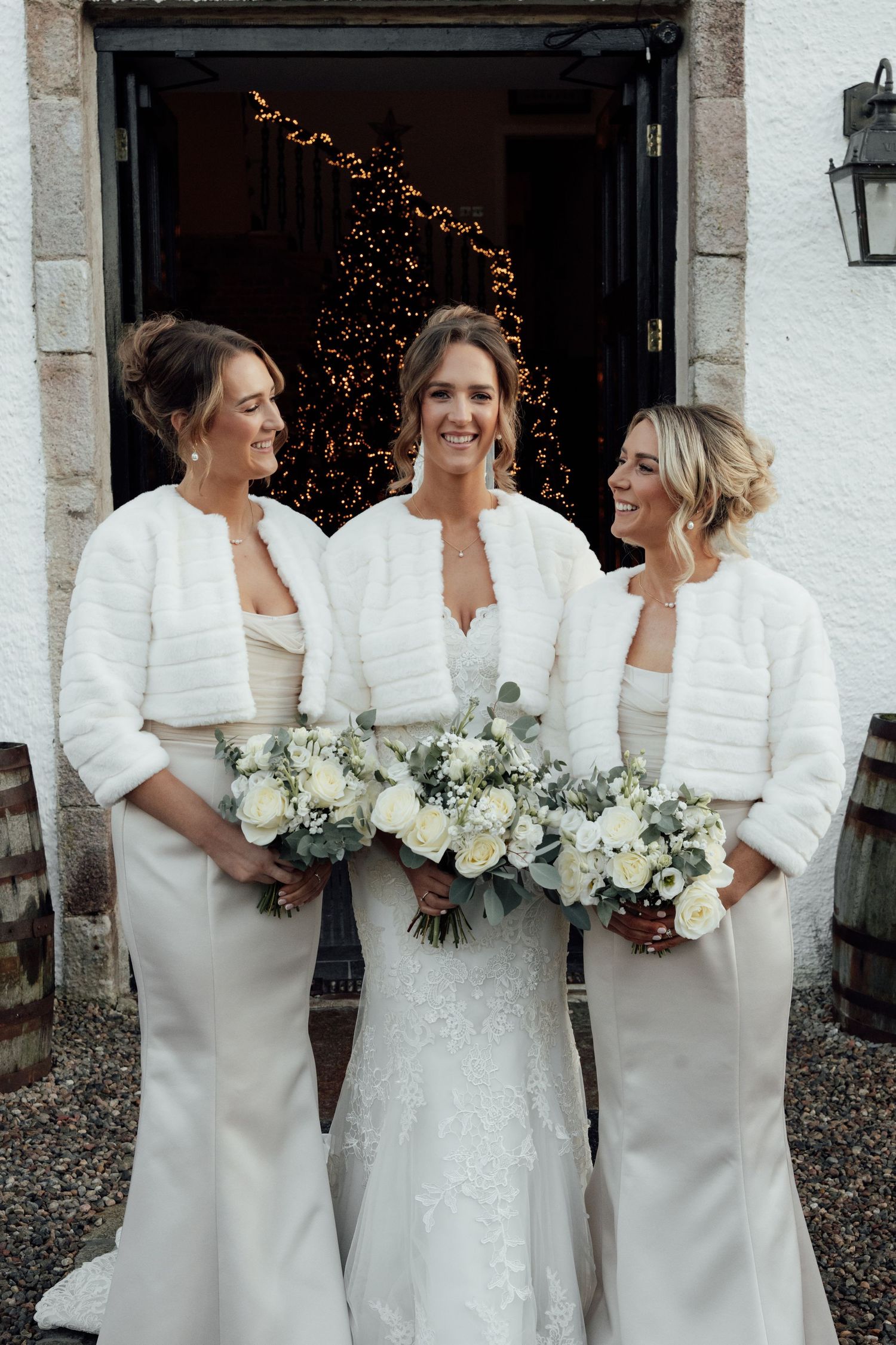 Wedding party members in coordinated white attire share laughs while holding matching white bouquets.