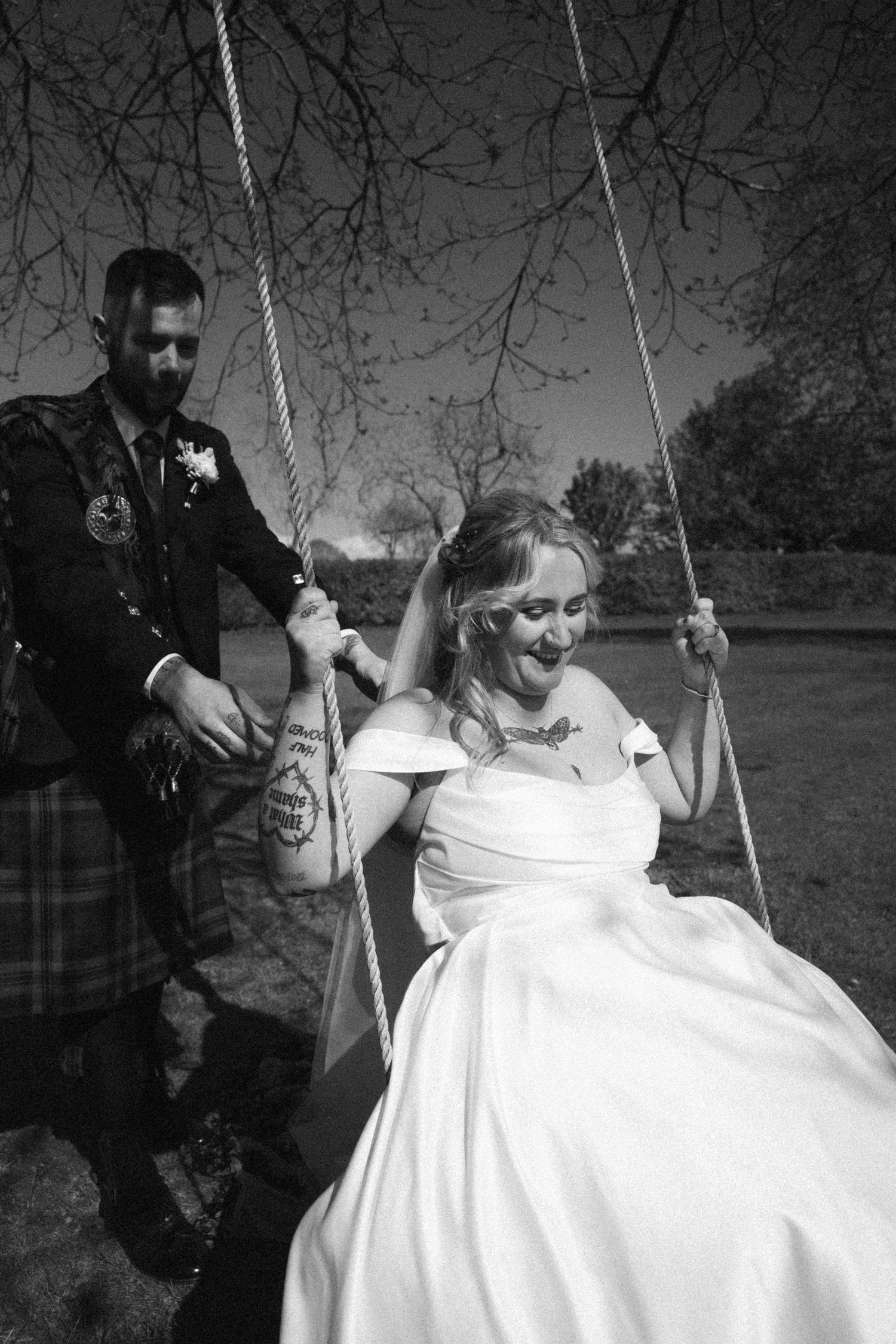 Groom in tartan kilt pushing laughing bride on rope swing in black and white wedding photo.