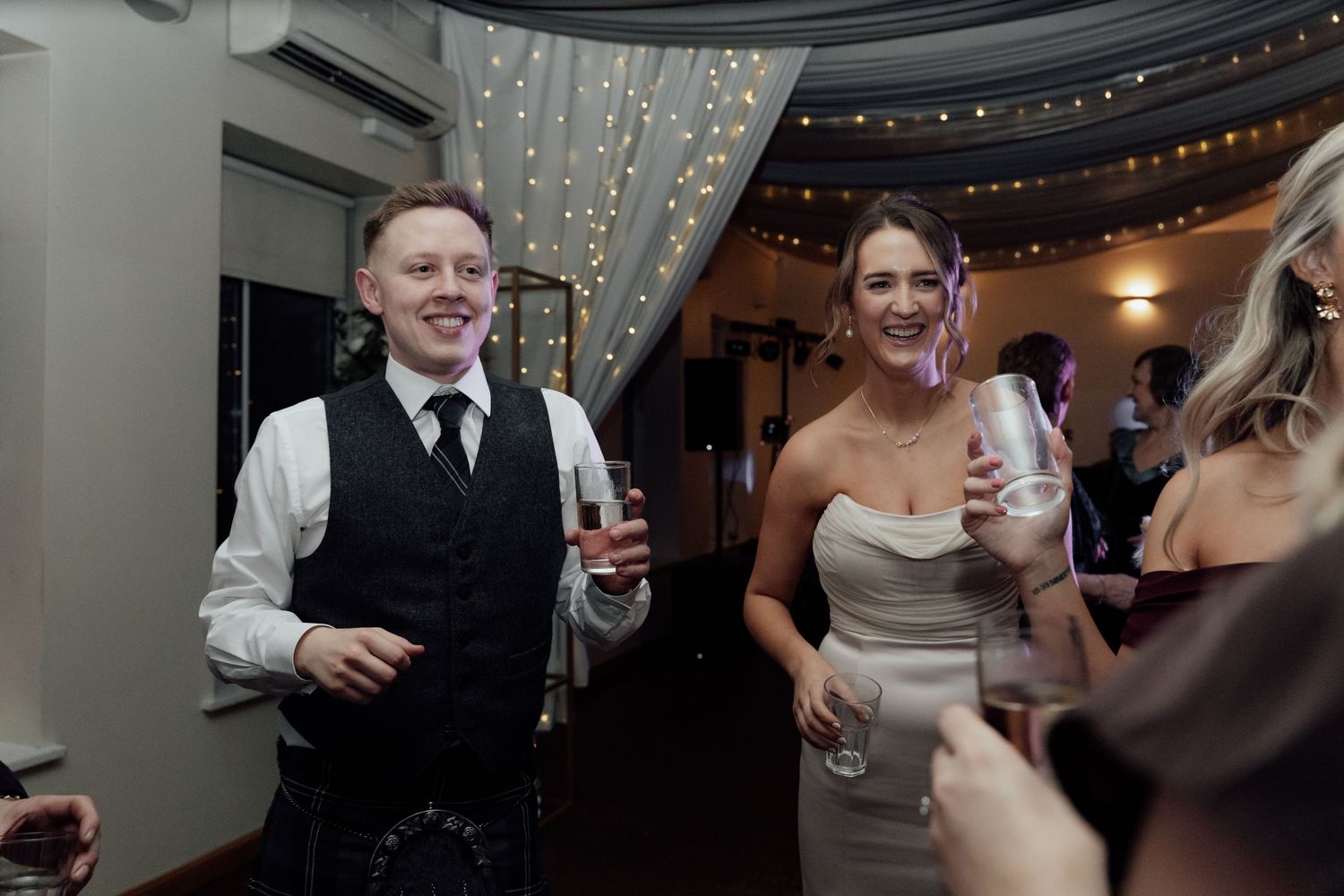 Wedding guests mingling and holding drinks under elegant draped ceiling with lights.