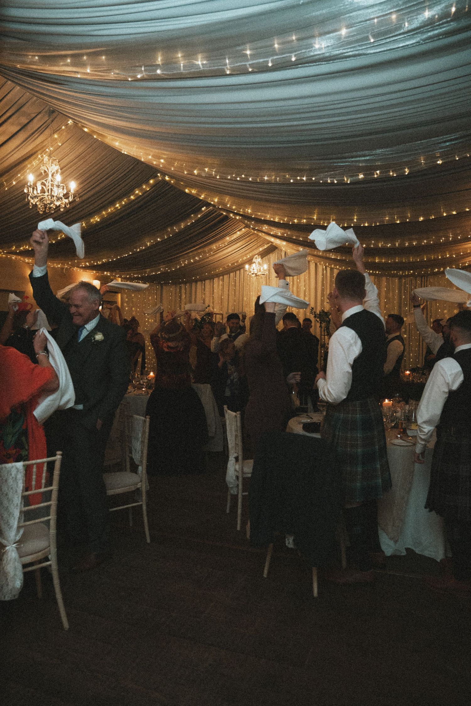 Guests enjoying a festive celebration under elegant draped ceiling with string lights and chandeliers.