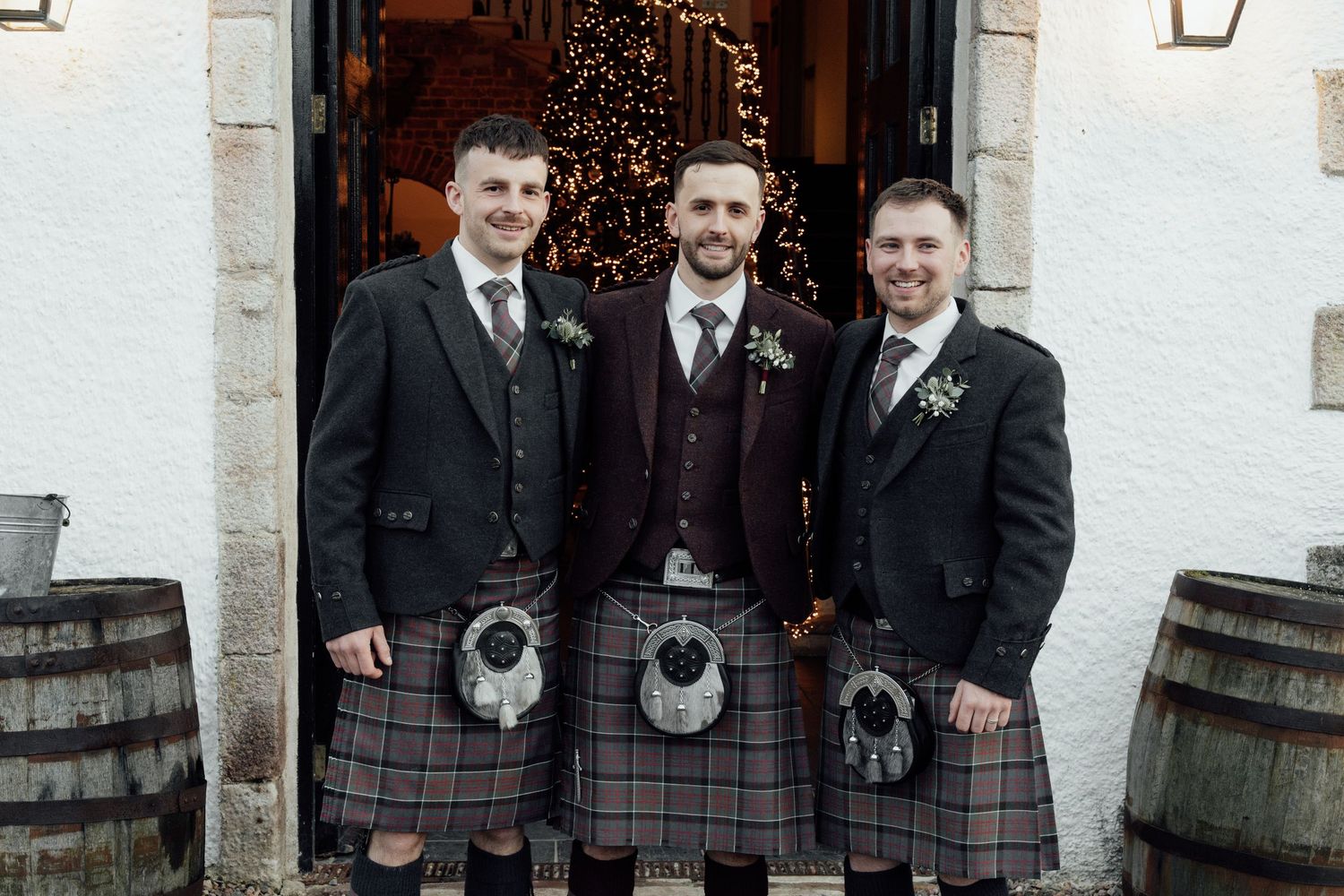 Groomsmen wearing matching kilts and dark jackets pose at church entrance with Christmas decor.