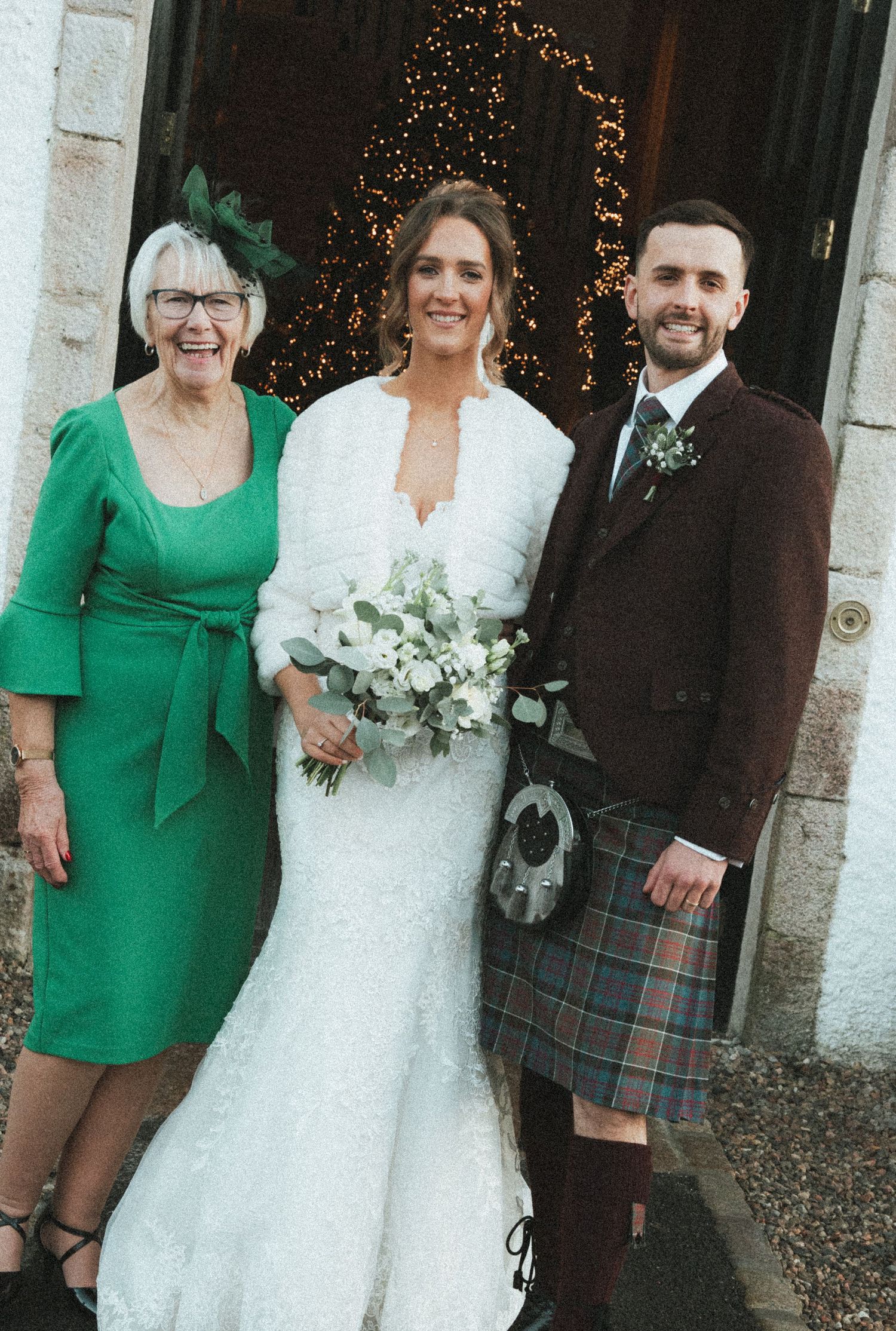 Wedding party poses outside stone building with bride in white dress and attendants in emerald green dresses.