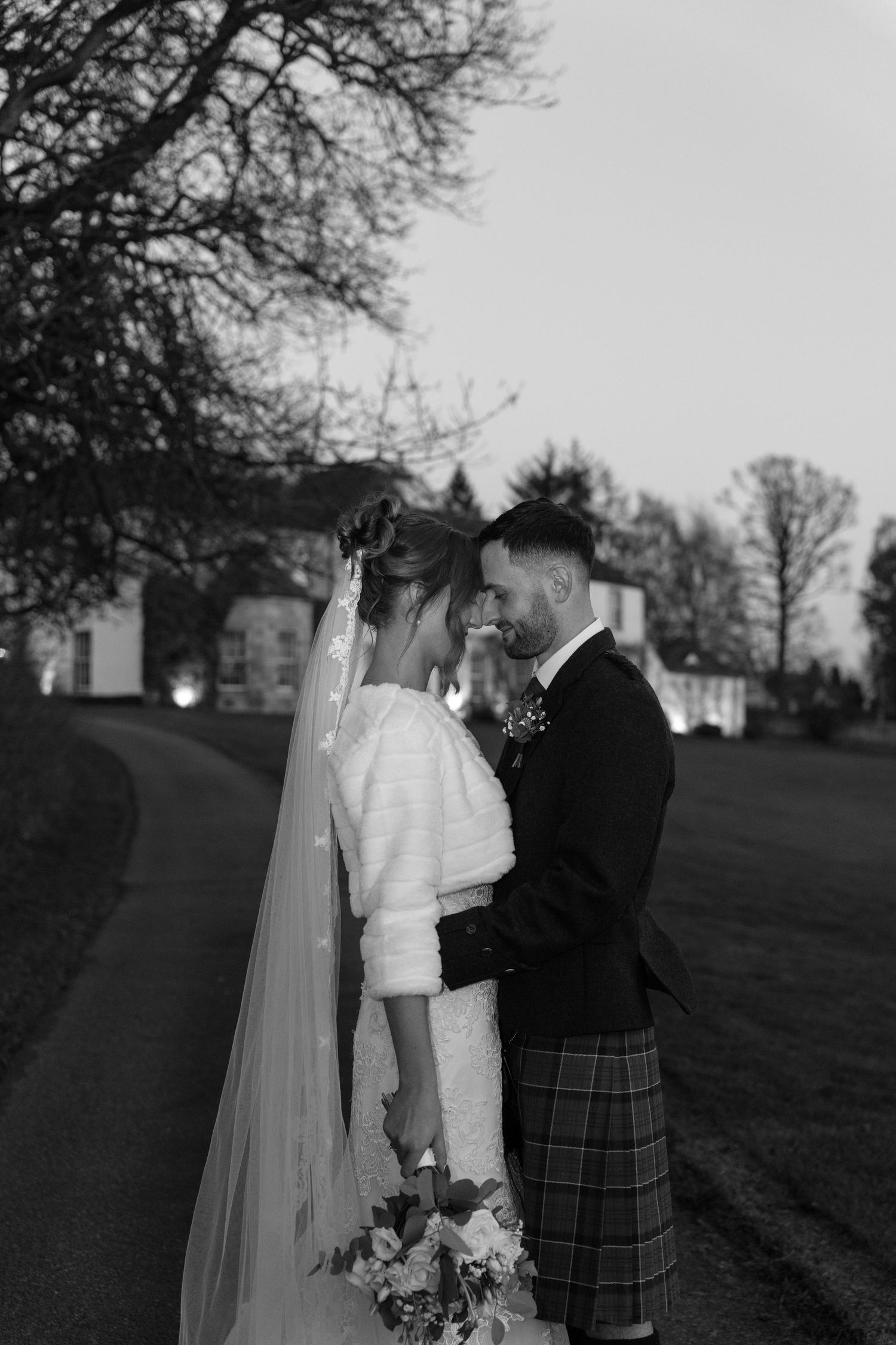 Black and white photo of newlyweds sharing a tender moment on a path near a grand estate.