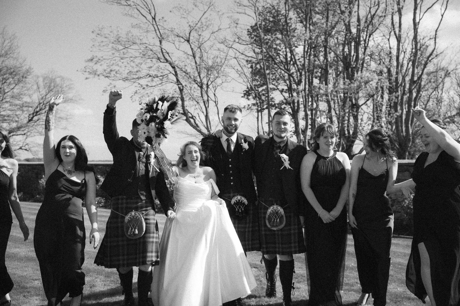 Black and white photo of wedding party celebrating outdoors, groom and groomsmen in kilts.