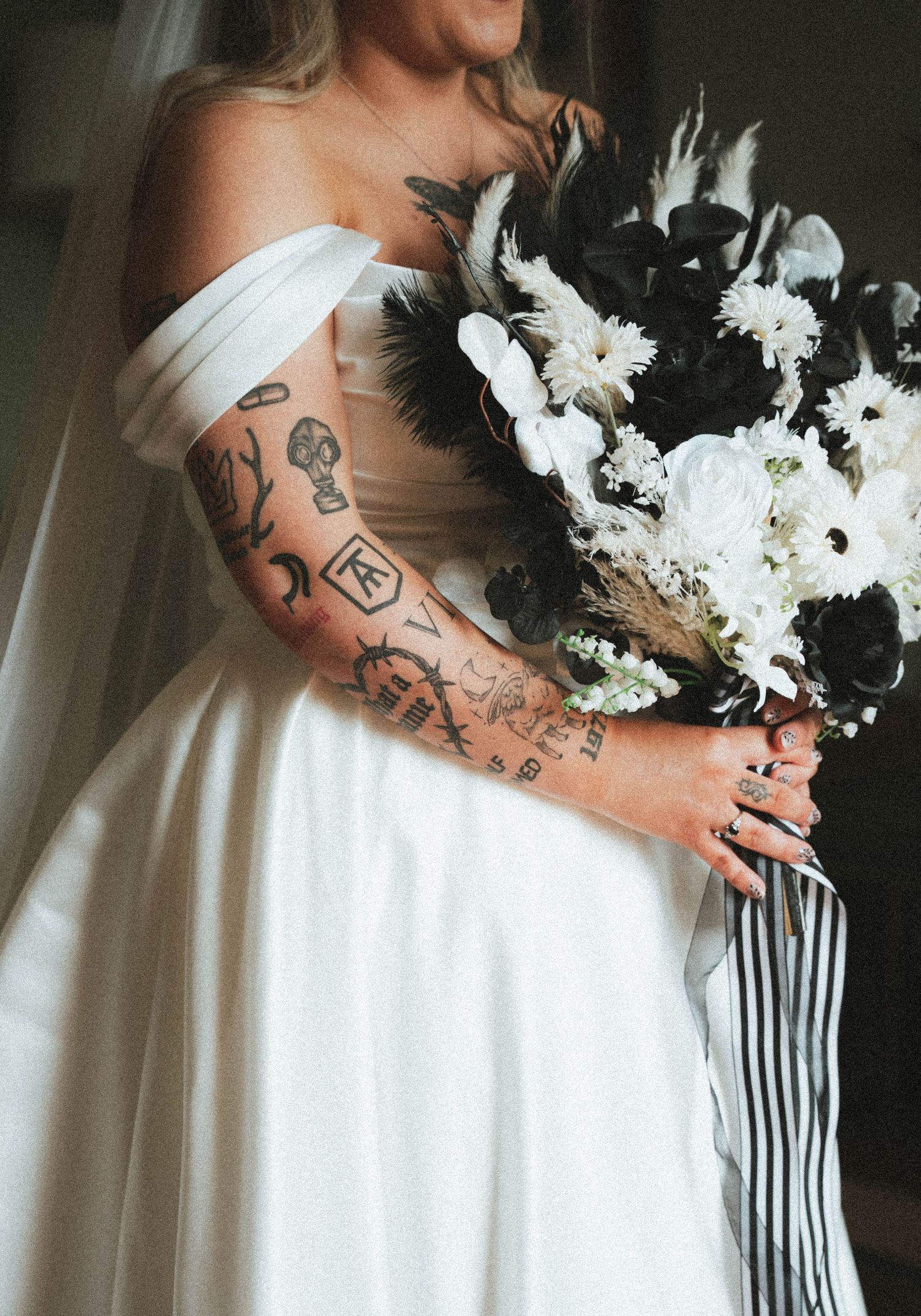 Tattooed bride in off-shoulder white gown holding a stunning black and white floral bouquet with feathers.