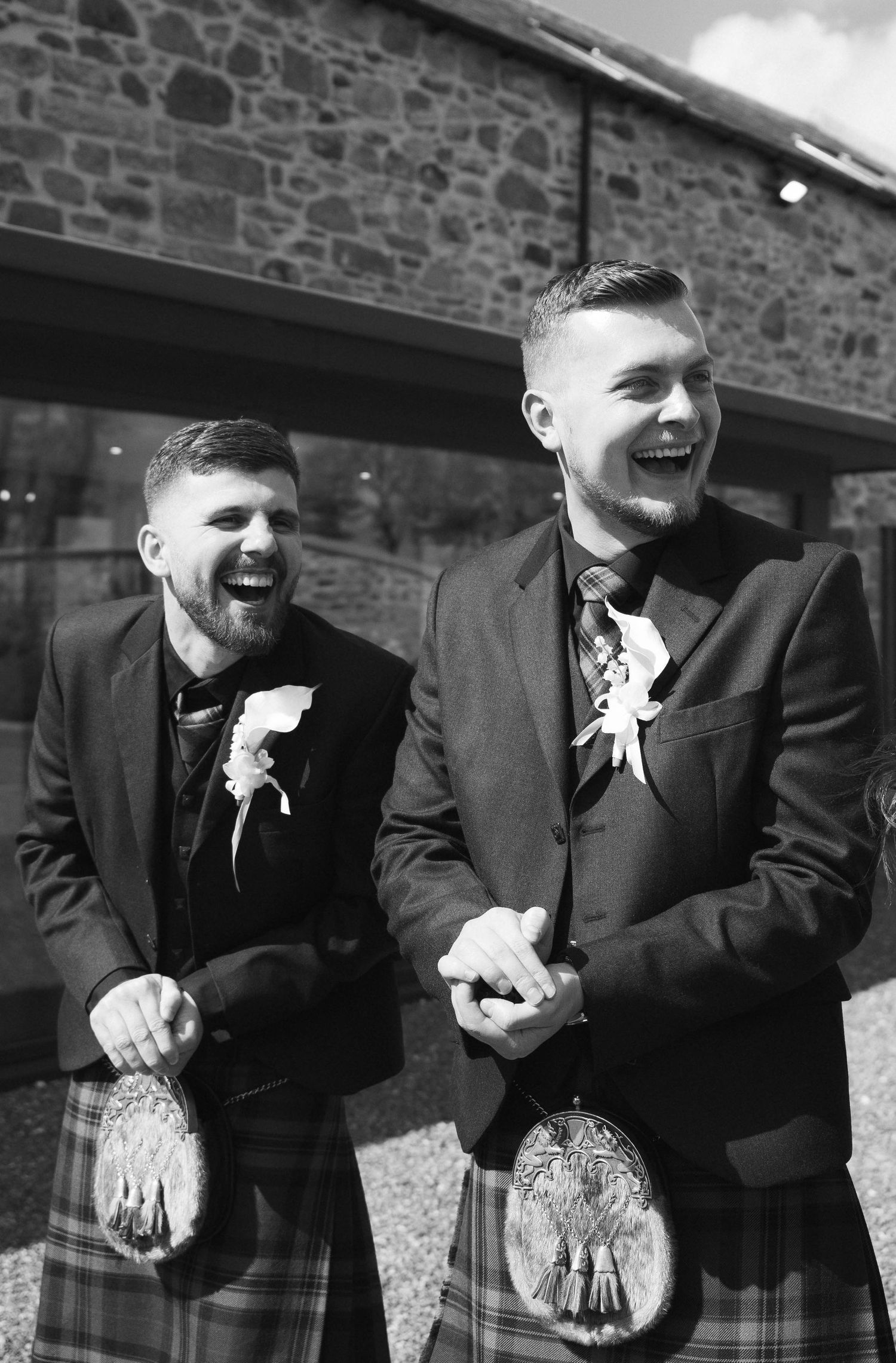 Two groomsmen in matching tartan kilts and boutonnieres laughing joyfully outside stone wedding venue.