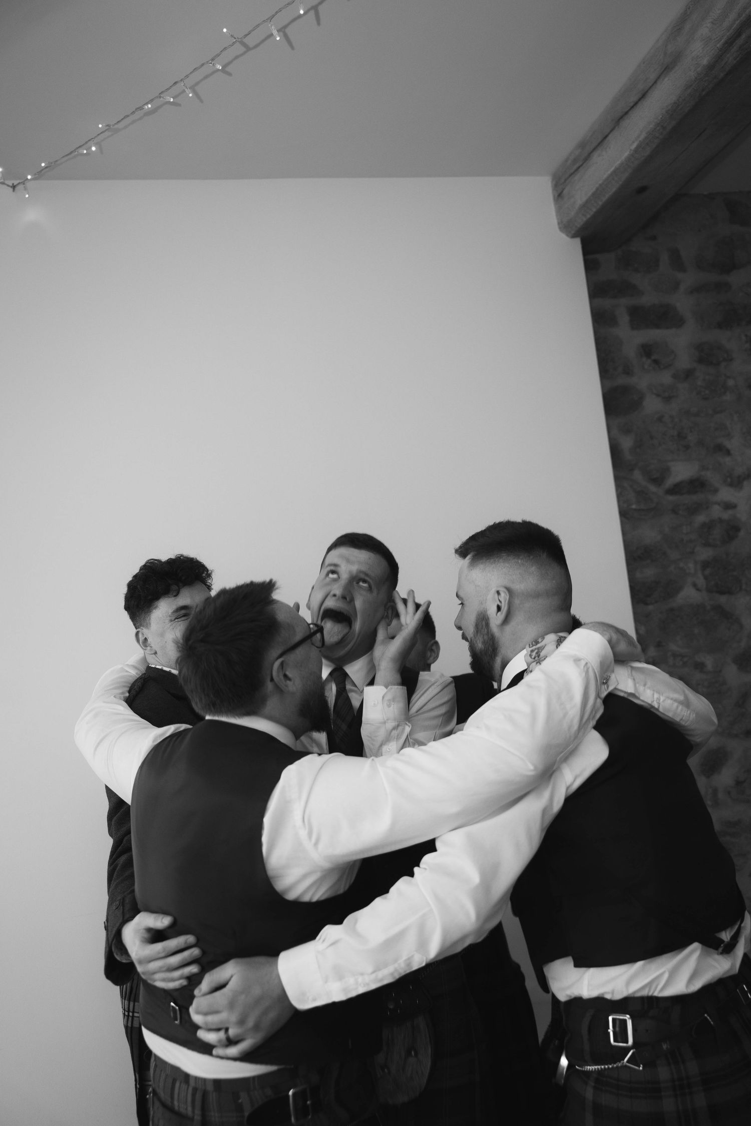 Groom laughs with groomsmen in kilts sharing a joyful candid moment against white wall at wedding.