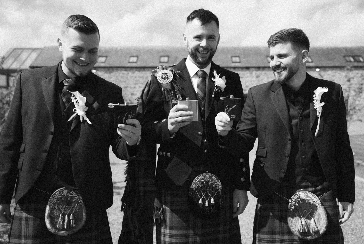 Three men in Scottish kilts and formal jackets laughing together at a wedding celebration outdoors.