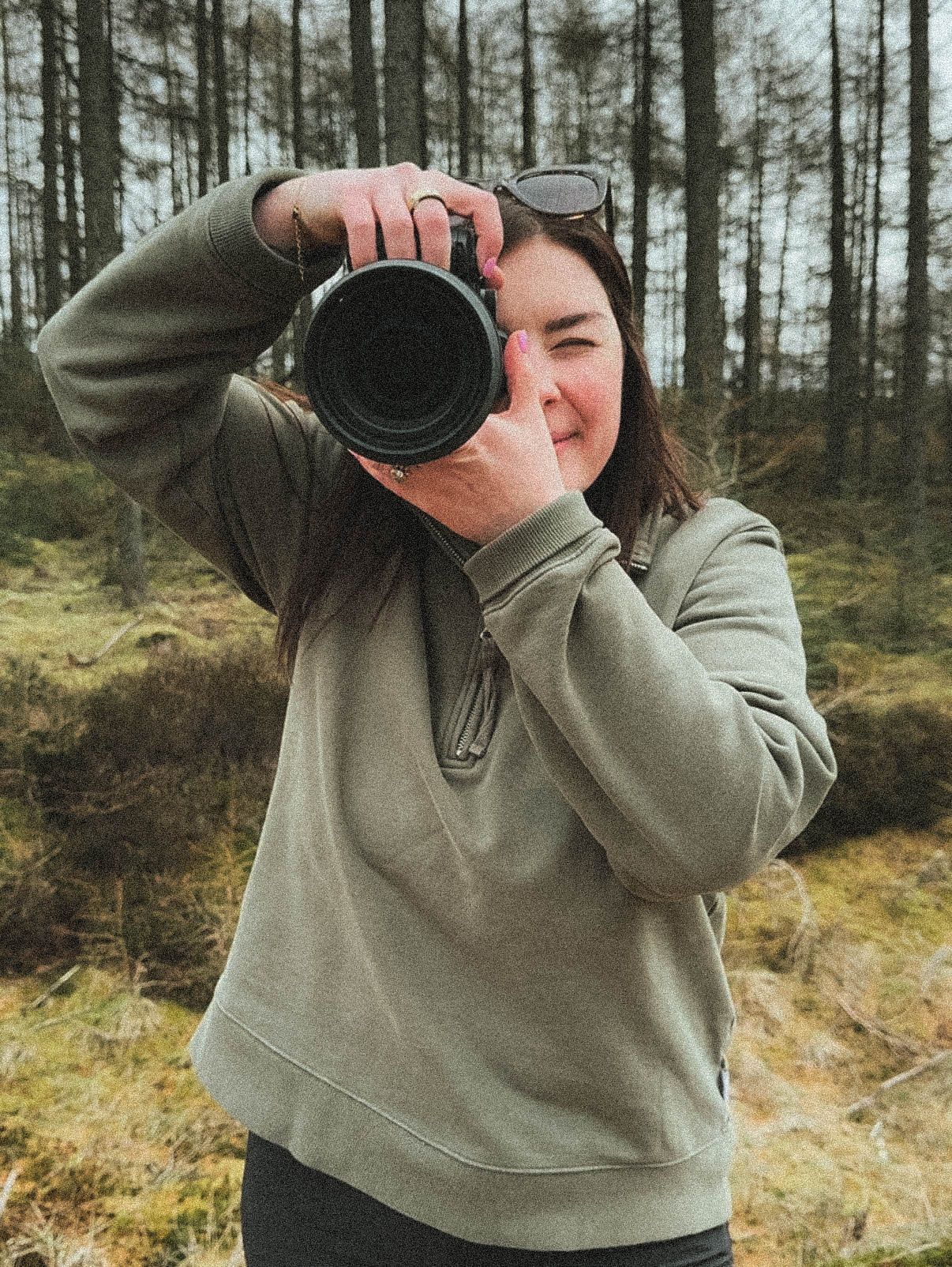 Woman in olive hoodie photographing in forest with DSLR camera, sunglasses on head, surrounded by trees.