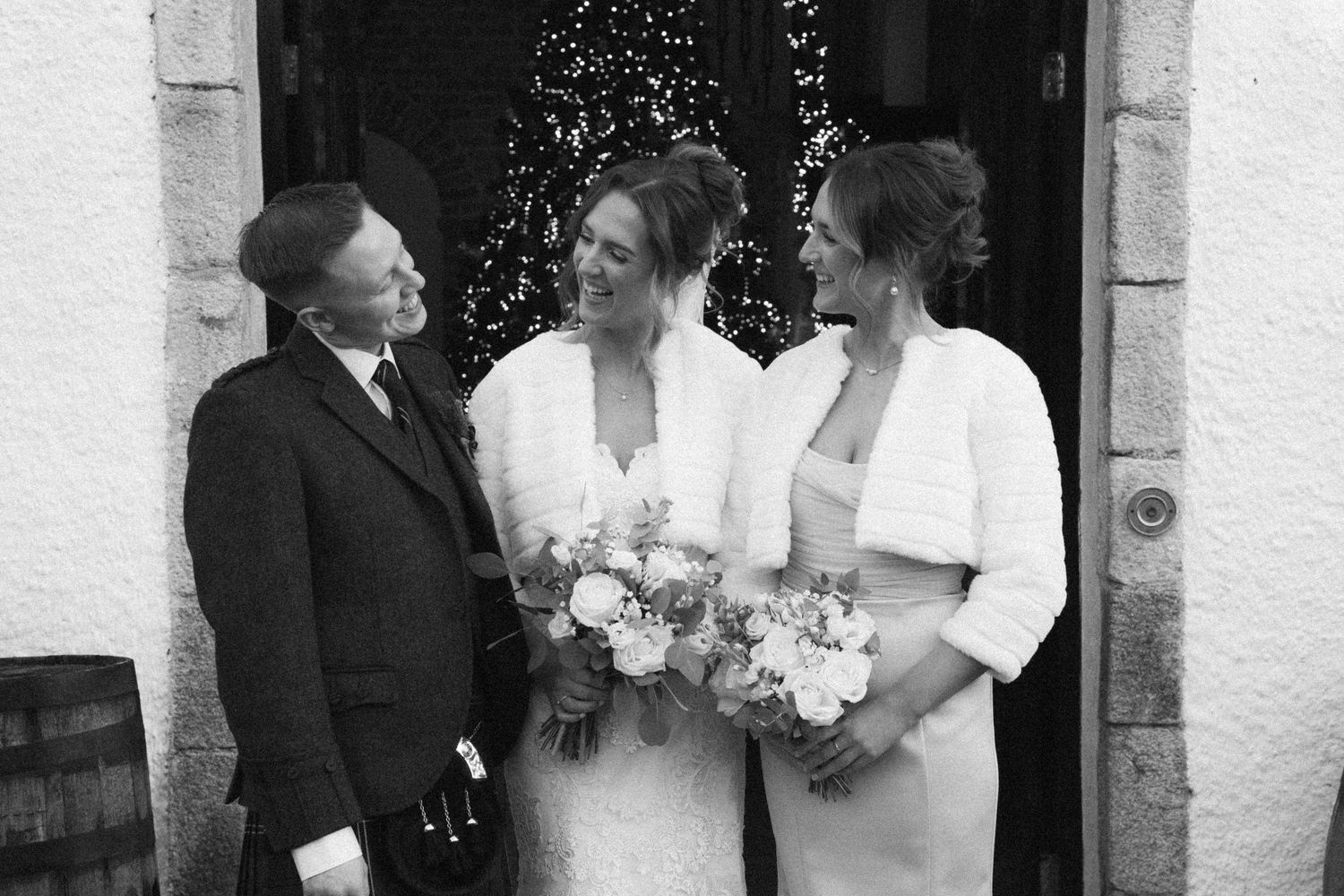 Wedding group portrait with elegant white dresses and bouquets against stone building entrance.