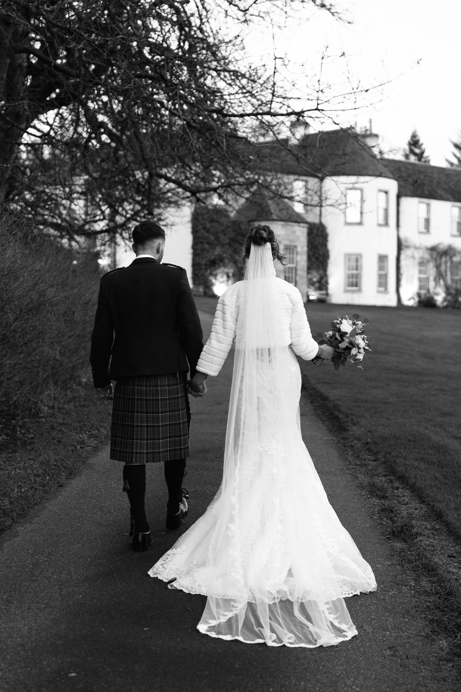 A couple walks along a path outside a grand white building, with the bride in a flowing white gown and groom in traditional kilt.