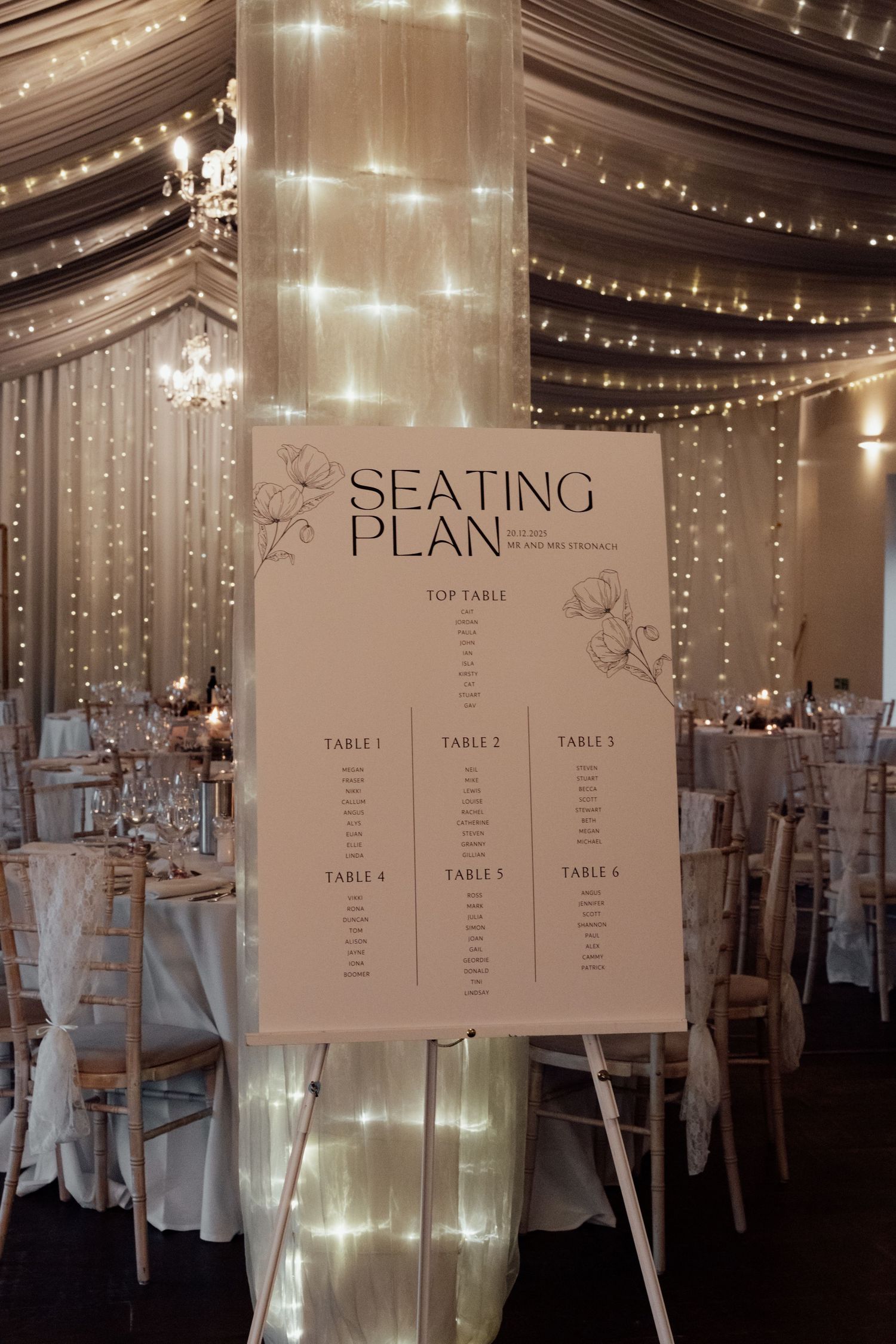 Elegant wedding reception seating plan display with fairy lights and white decor.