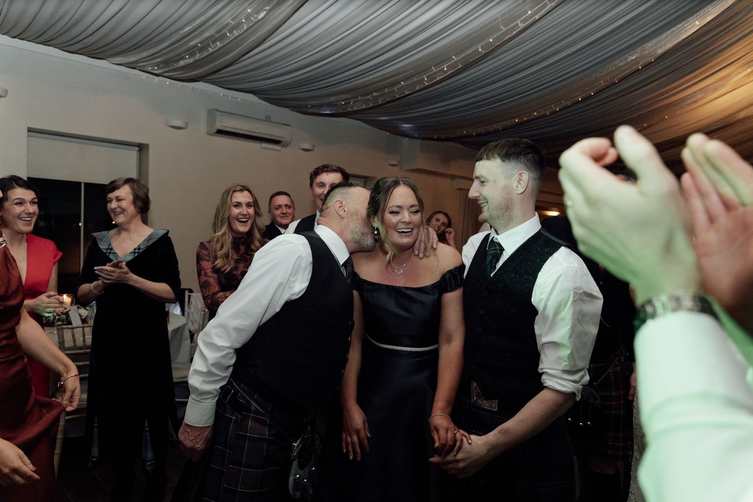 Lively dance floor moments captured at wedding reception under draped fabric ceiling.