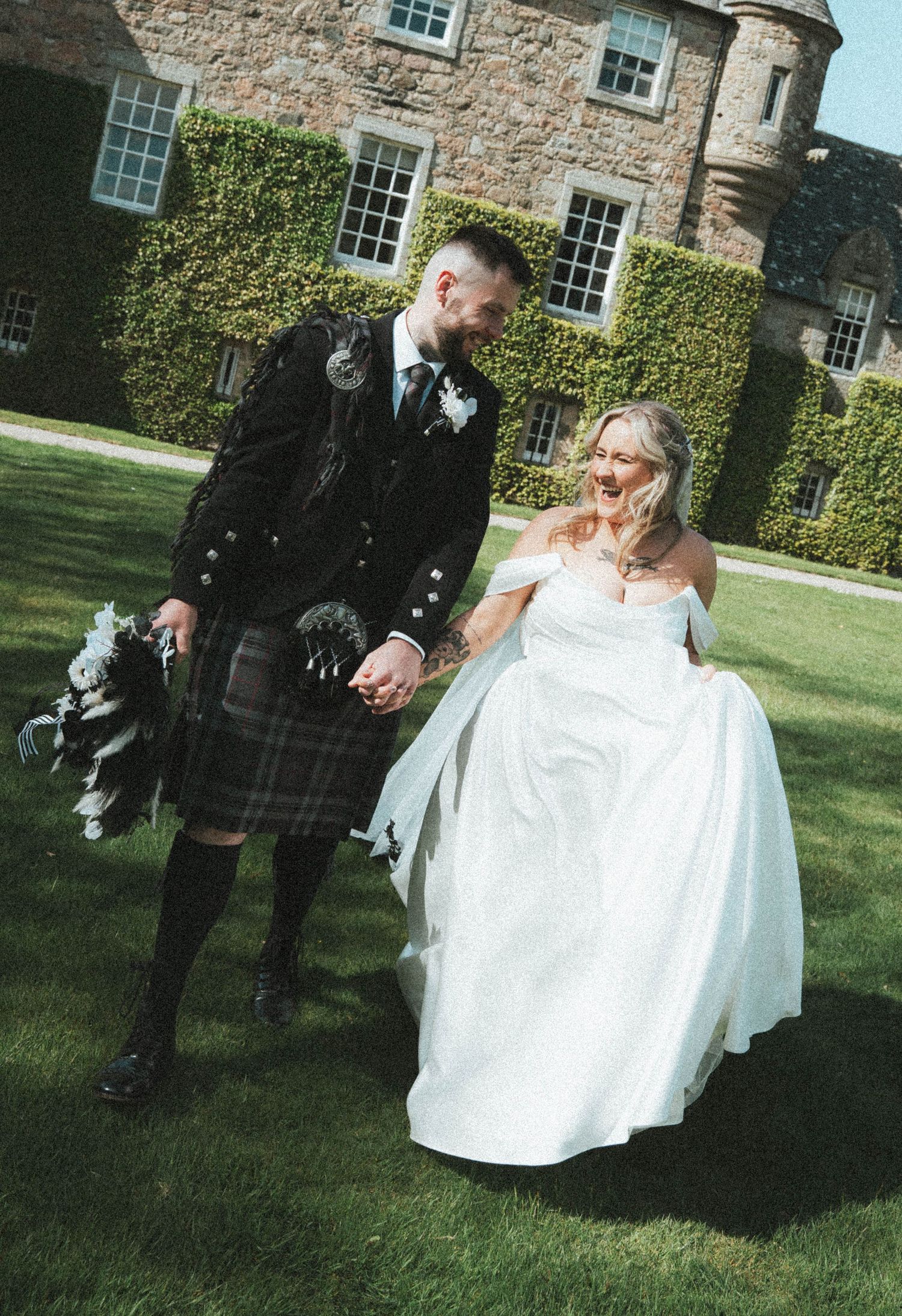 Groom in Scottish kilt and bride in white gown walking hand in hand outdoors.