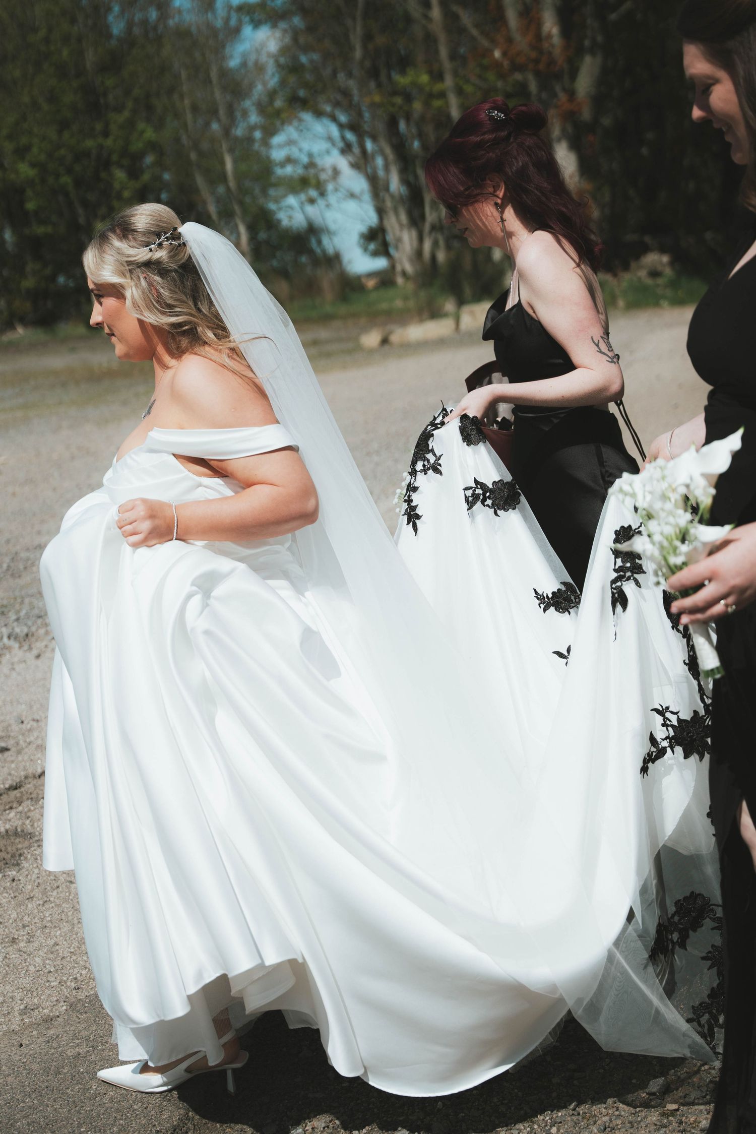 Bride in white off-shoulder gown with veil, assisted by bridesmaid in black dress outdoors.
