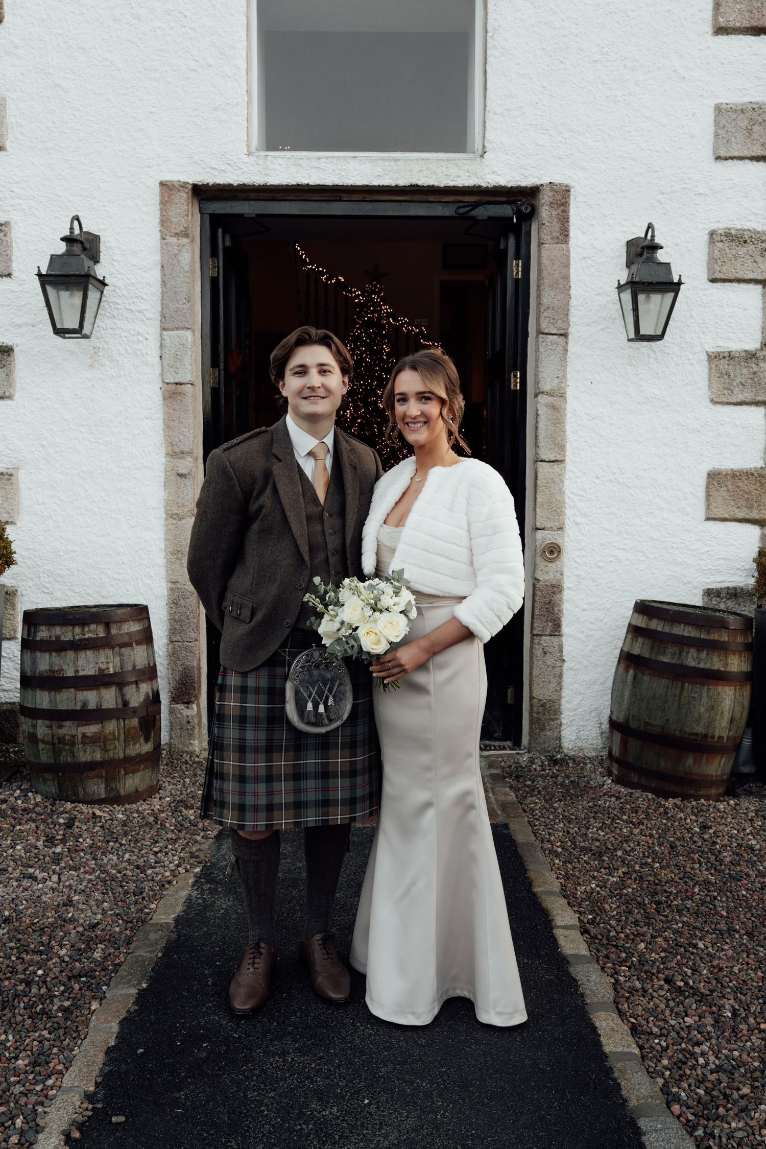 Wedding couple poses outside stone building with traditional Scottish and modern formal attire.