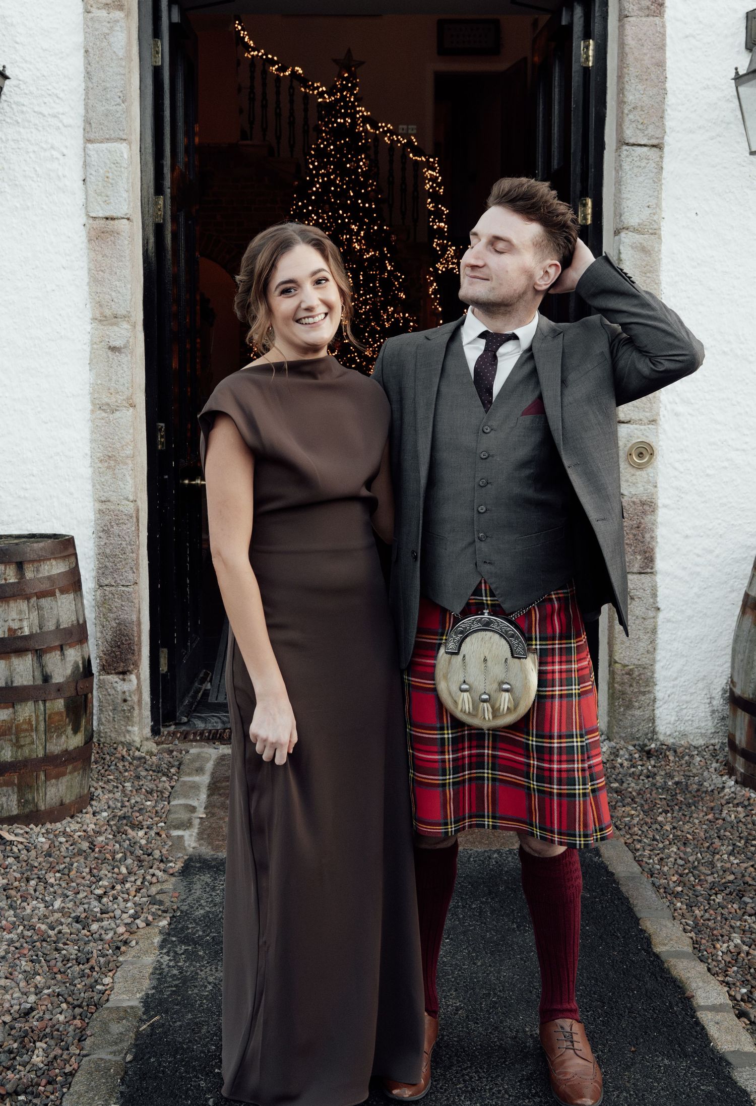 A couple shares a lighthearted moment in formal attire, with the man wearing a traditional red tartan kilt.