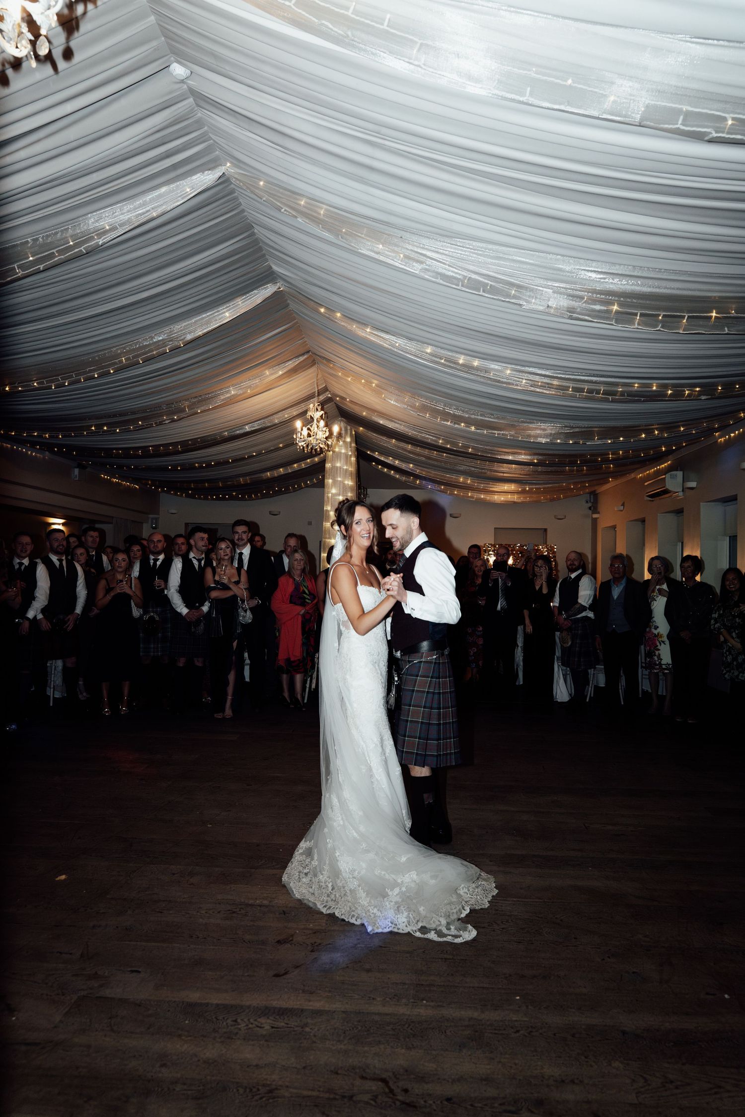 Couple dancing in a white tent reception venue decorated with twinkling lights and chandeliers.