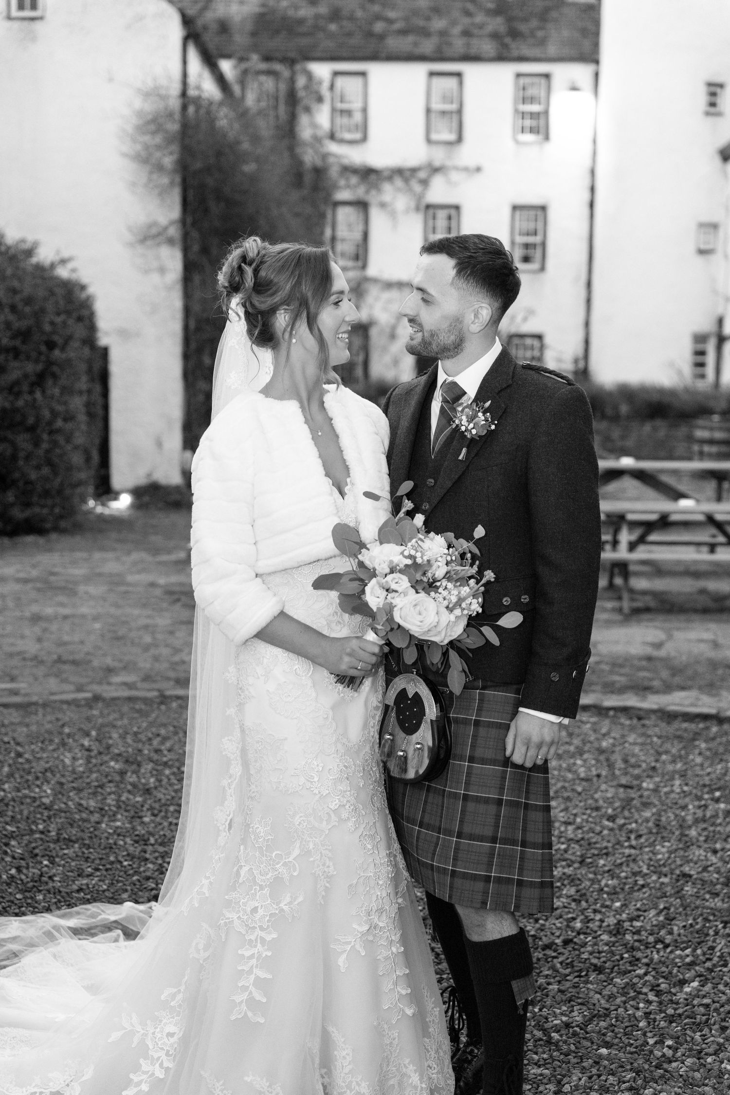 A romantic wedding photo in black and white showing a couple in traditional Scottish attire outside a historic building.