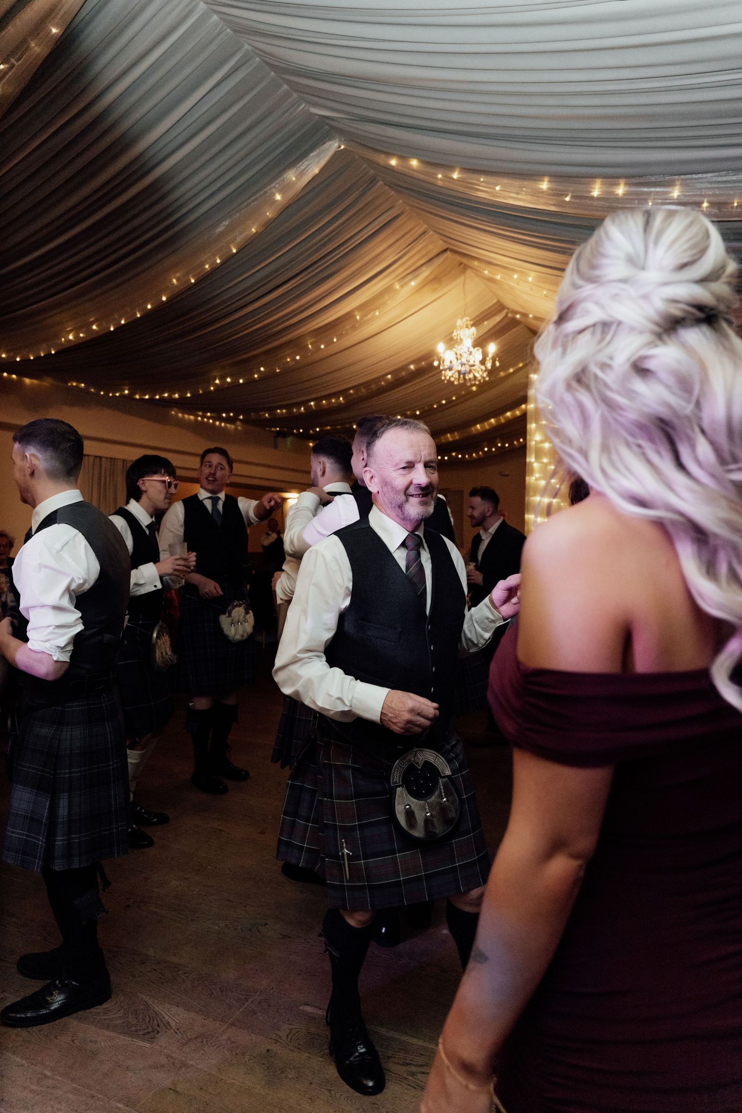 Wedding reception dance floor with Scottish kilts and formal attire under romantic lighting.