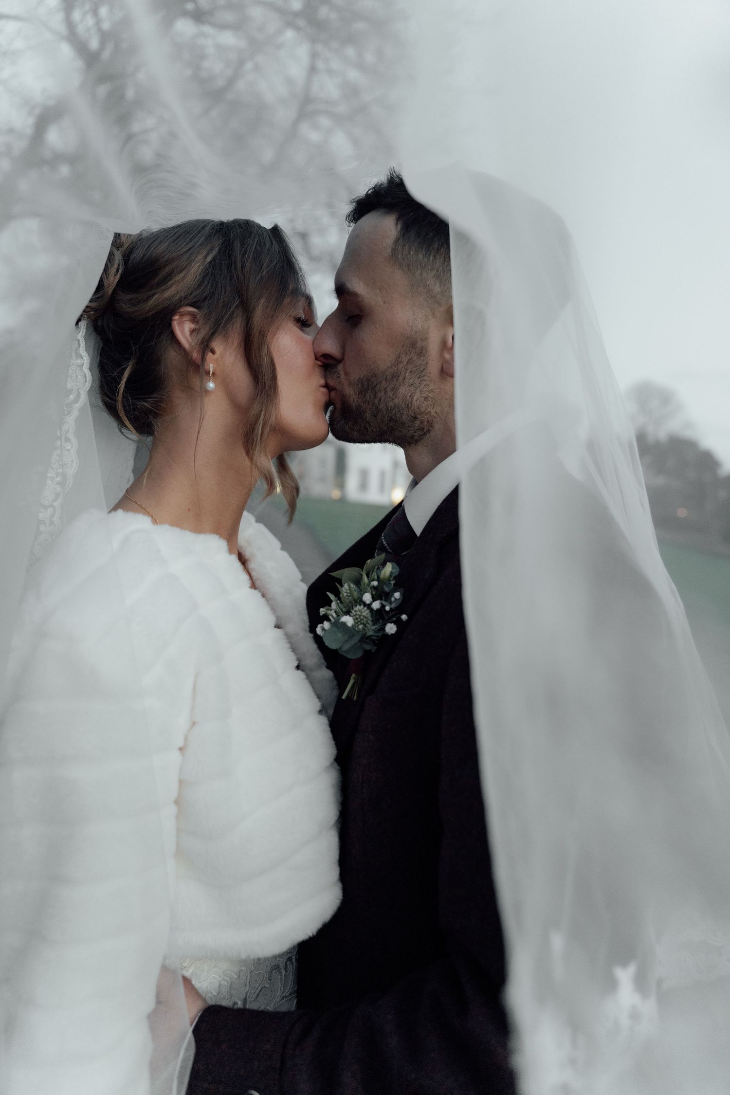 Newlyweds embrace in a tender moment beneath a sheer veil during their outdoor wedding photoshoot.