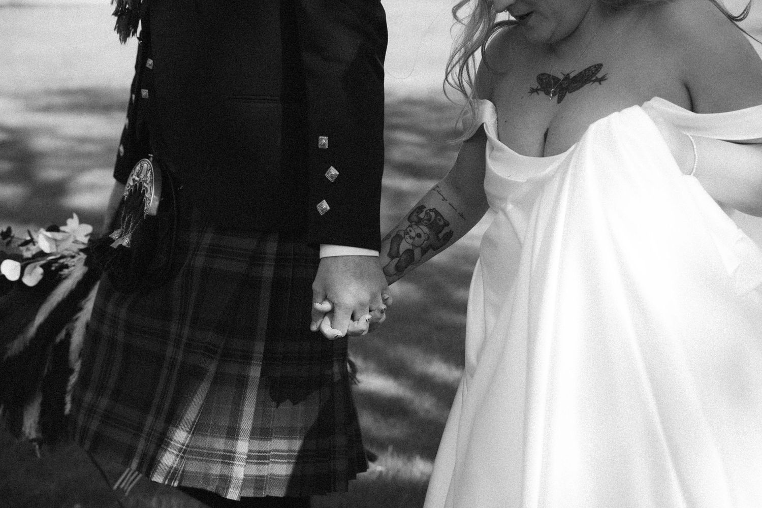 Black and white close-up of bride and kilted groom holding hands showing tattoos.