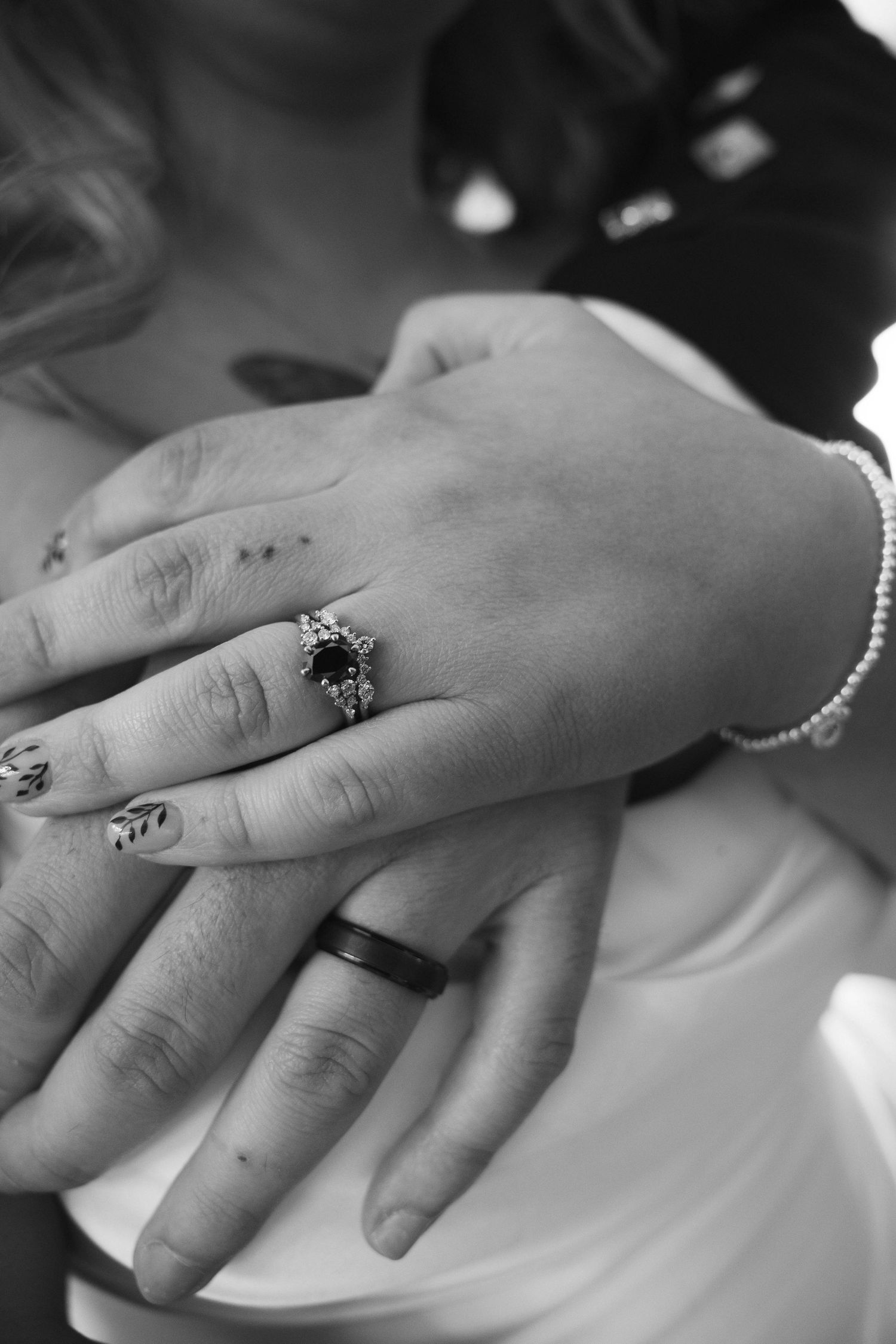 Close-up black and white photo of newlyweds hands showing dark gemstone engagement ring and black wedding band.