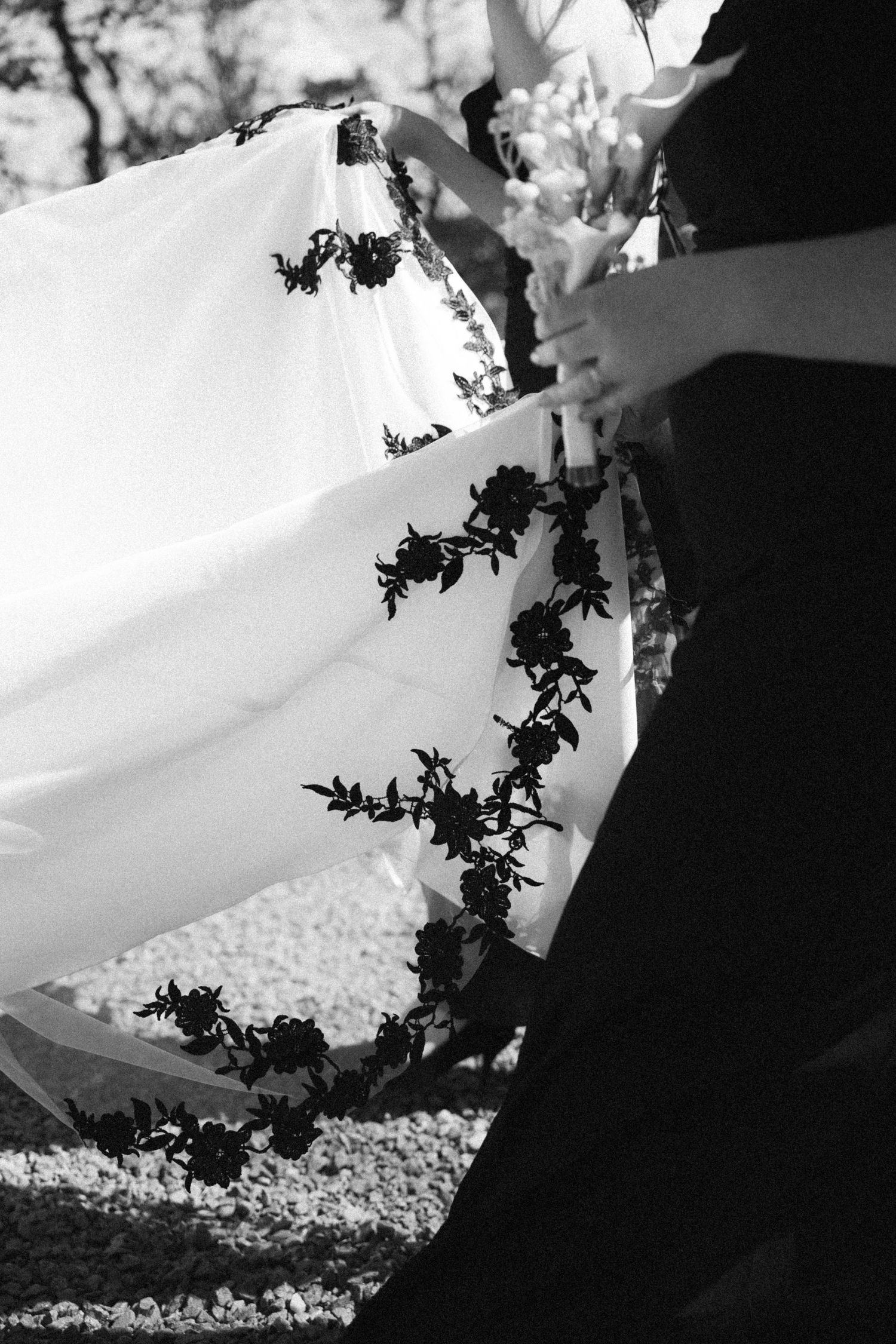 Black and white close-up of bride's wedding veil with delicate floral embroidery details being held by attendants.