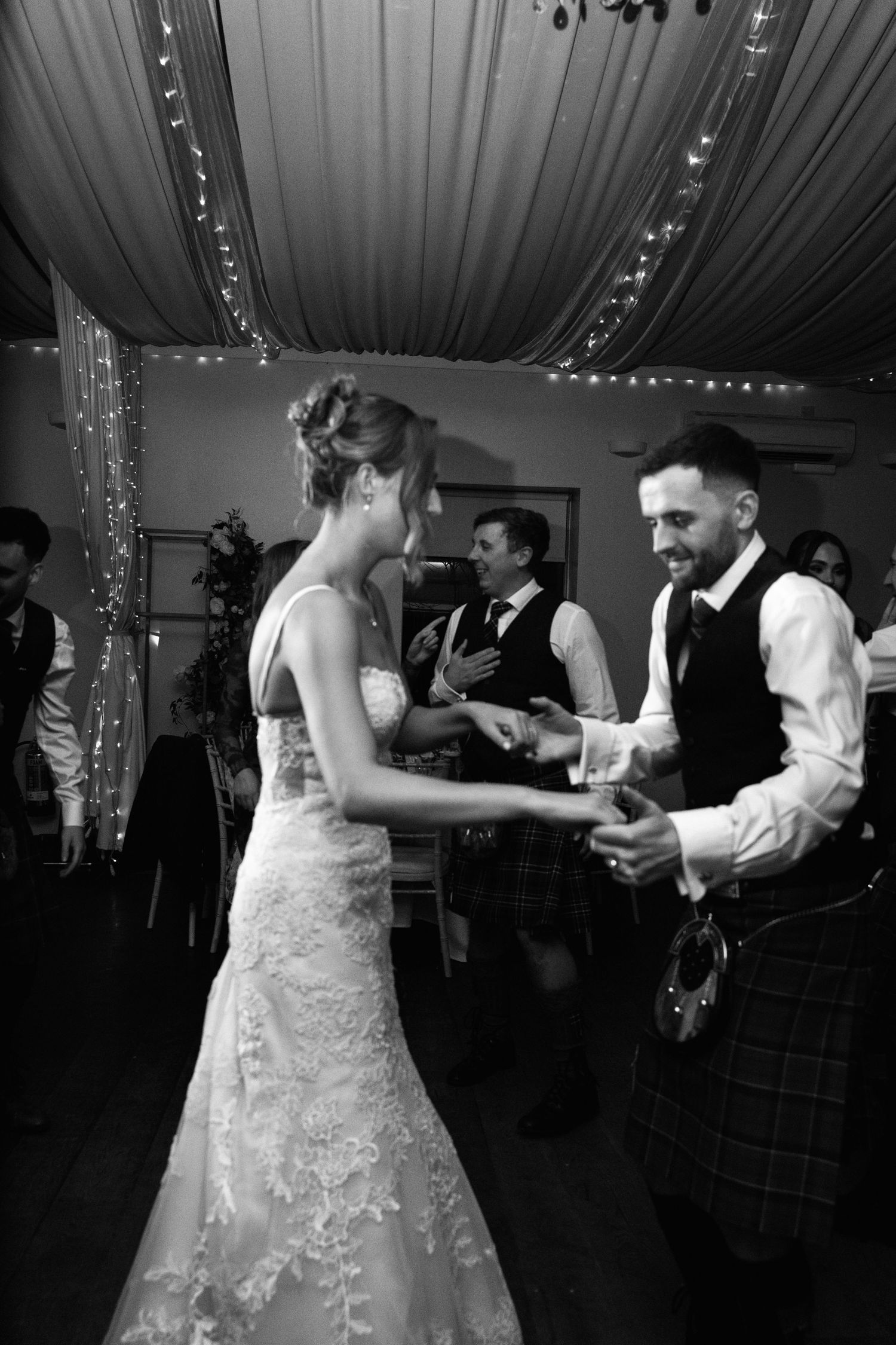 Black and white photo of wedding couple dancing at reception under draped ceiling with string lights.