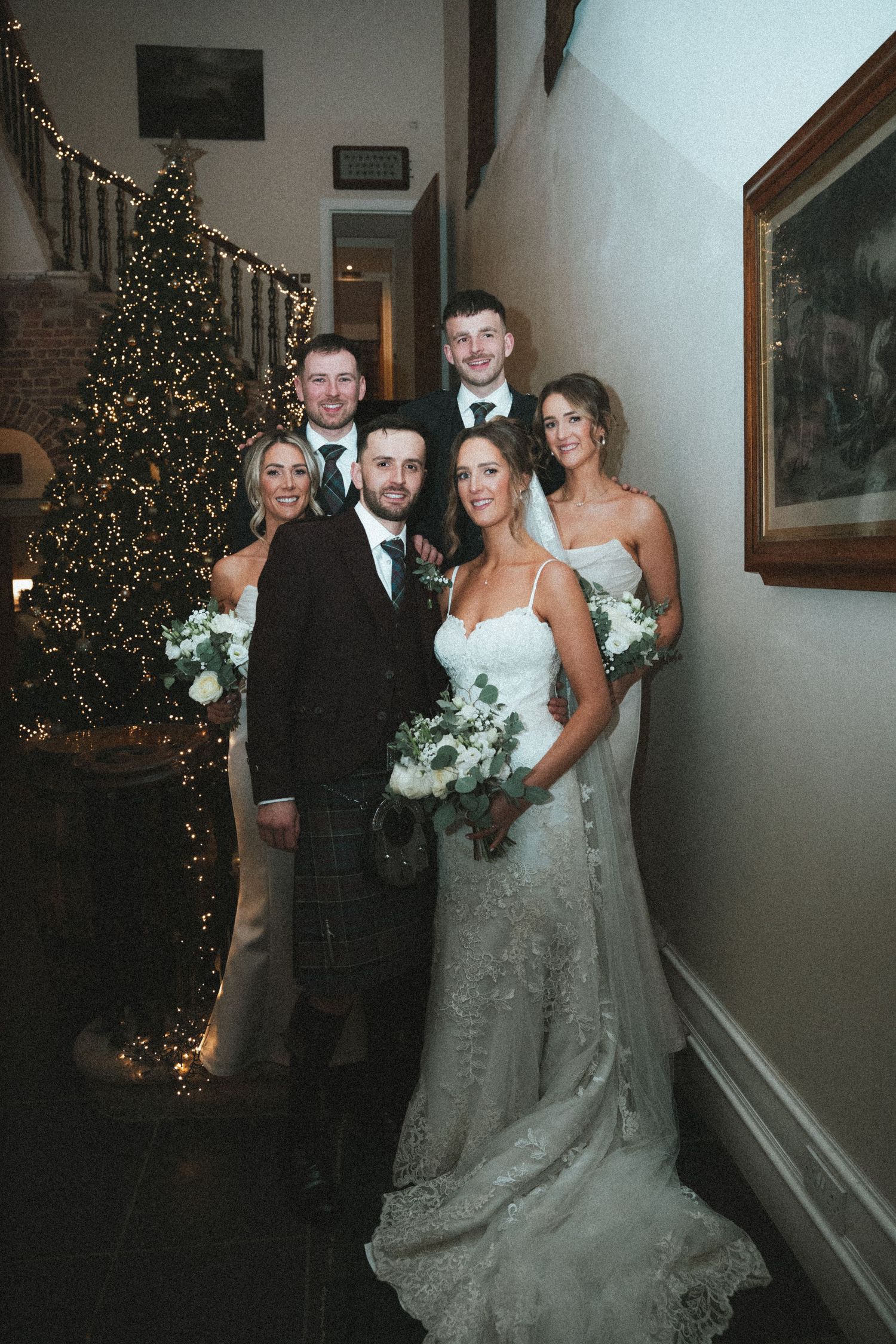 Wedding party poses in front of decorated Christmas tree in elegant indoor venue with white walls and artwork.