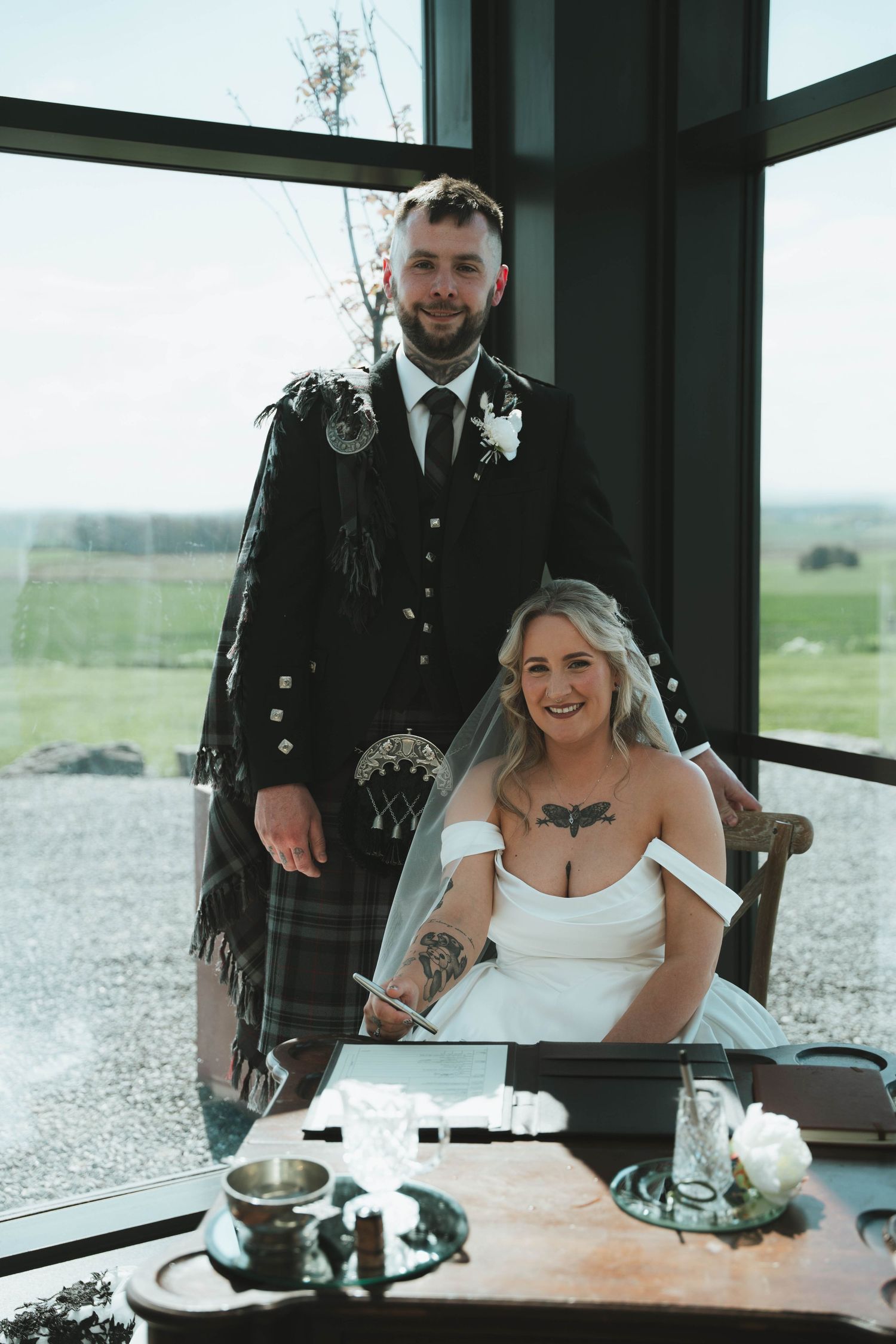 Groom in Scottish kilt standing beside seated bride in white dress signing wedding register at table by window.