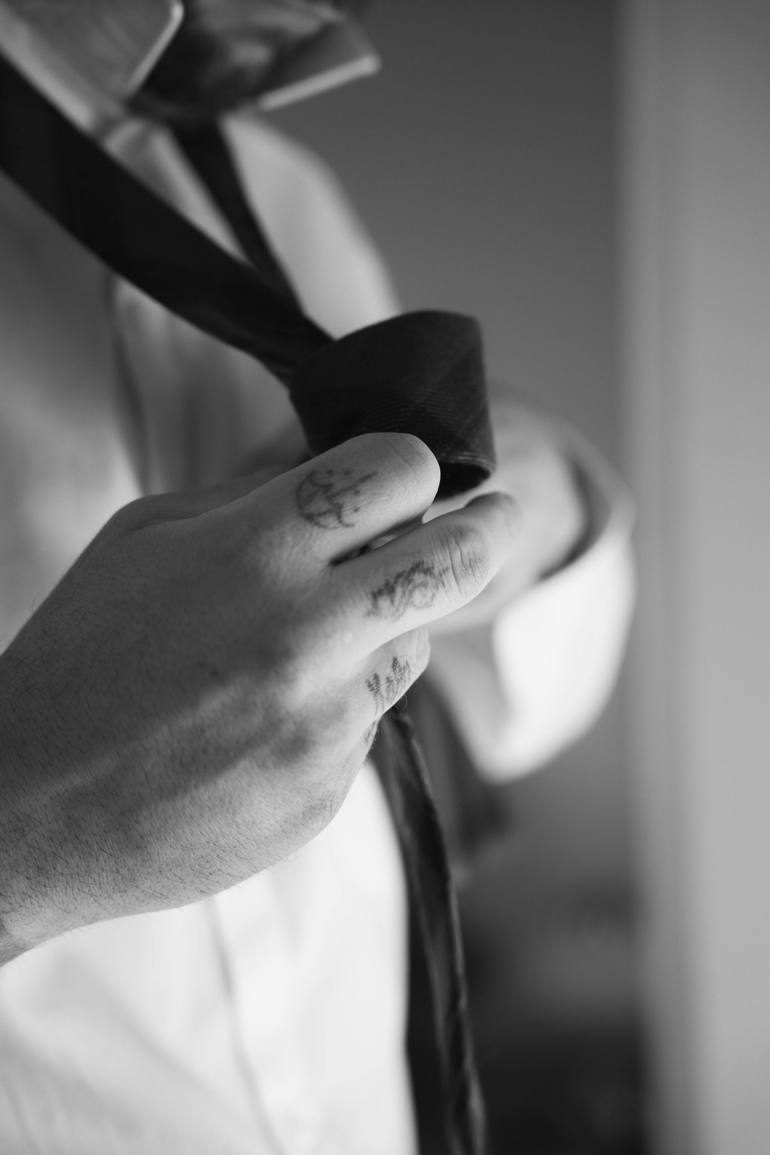 Close-up black and white photo of a tattooed groom adjusting his tie before the wedding ceremony.