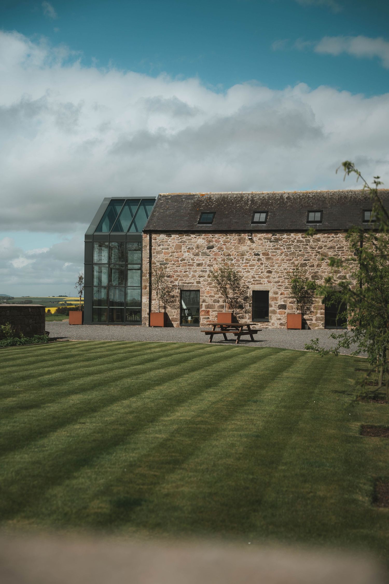 Stone barn wedding venue with modern glass extension, striped lawn, and picnic bench under a blue sky.