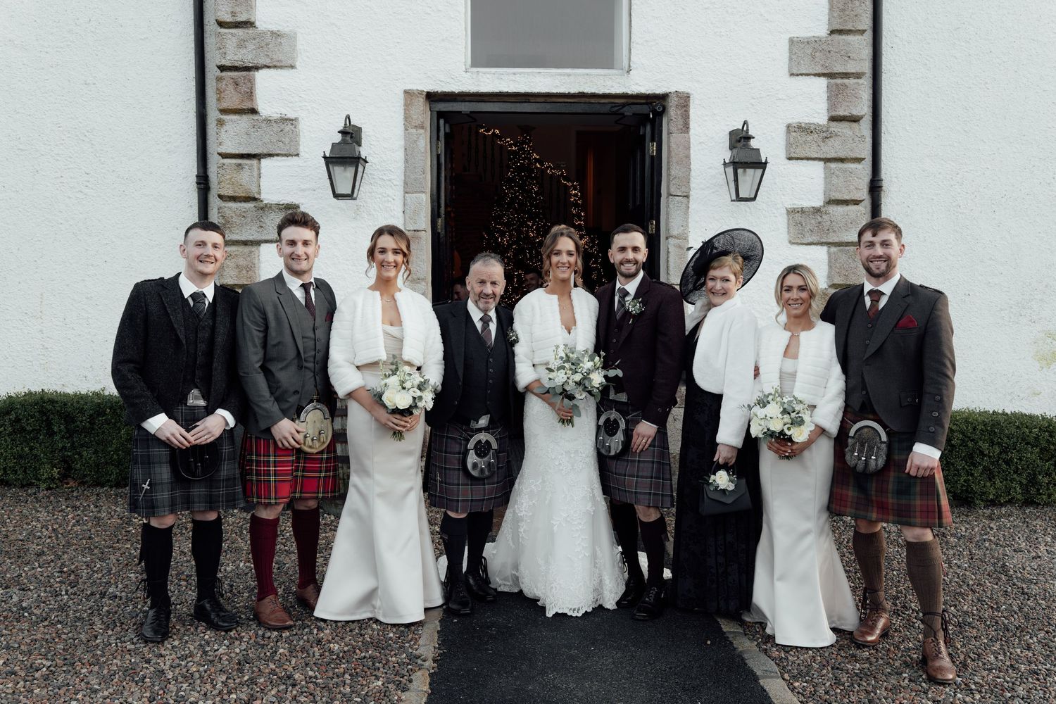 Wedding party gathered outside white stone building with traditional lanterns and Scottish kilts.