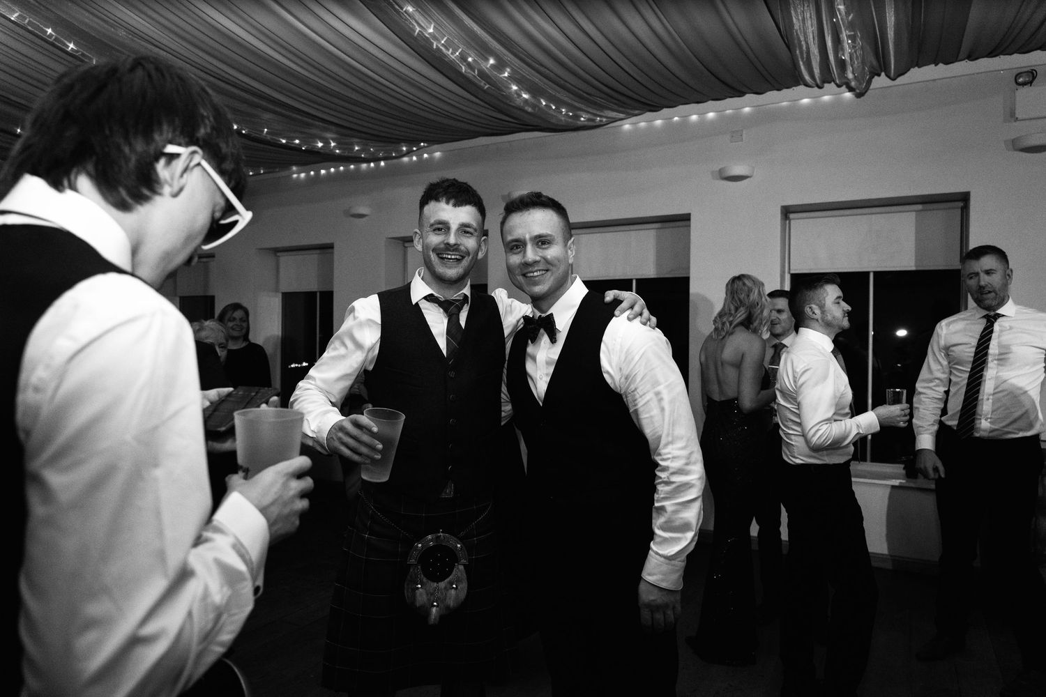 Black and white photo of guests in kilts and formal wear at a Scottish-themed wedding reception.