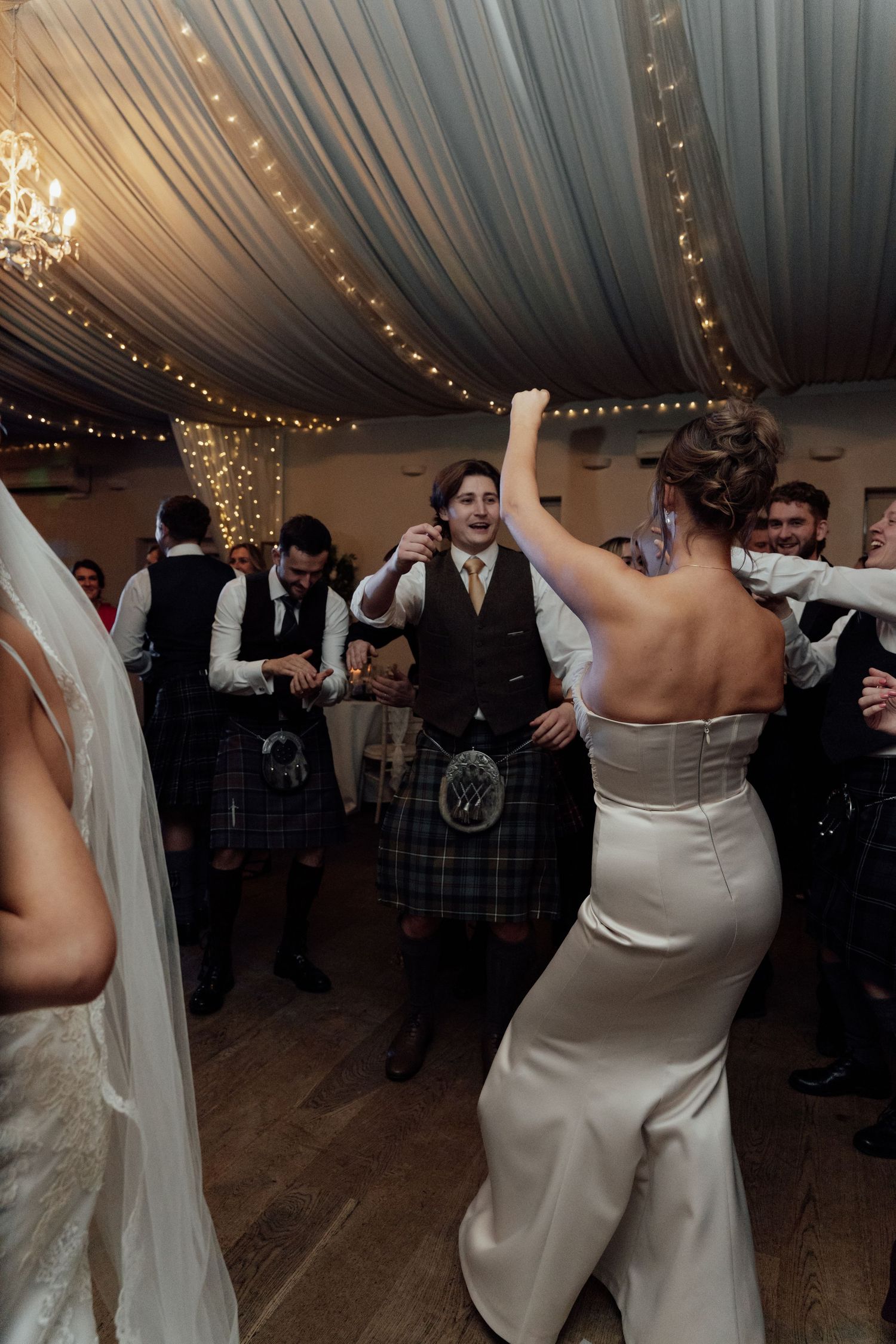 Wedding reception dance floor filled with celebration under elegant draped ceiling and chandeliers.